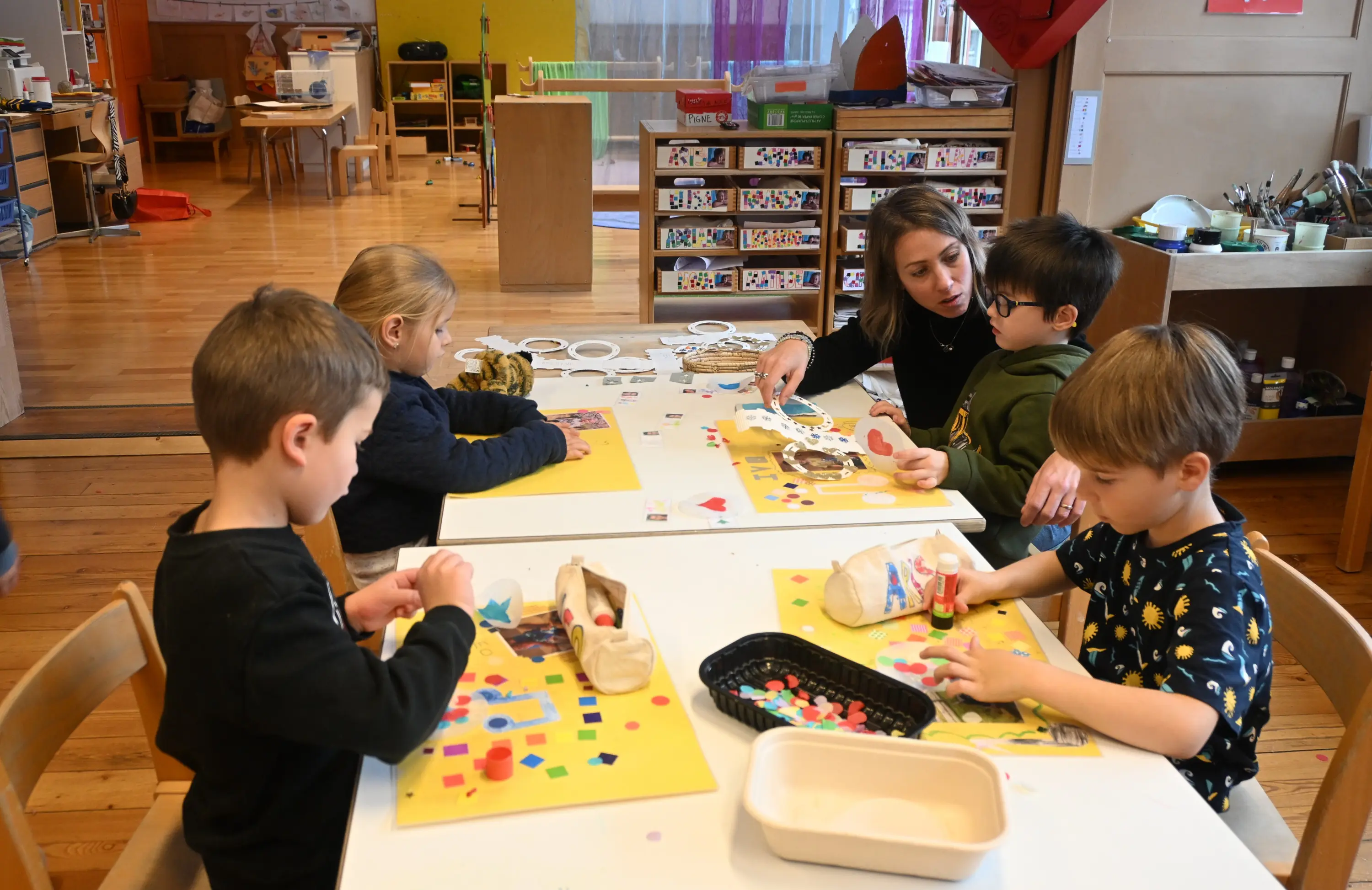 Un gruppo di bambini sta creando artigianato su tavoli di lavoro, mentre un insegnante li guida. I bambini utilizzano materiali colorati come cartoncini e adesivi. L