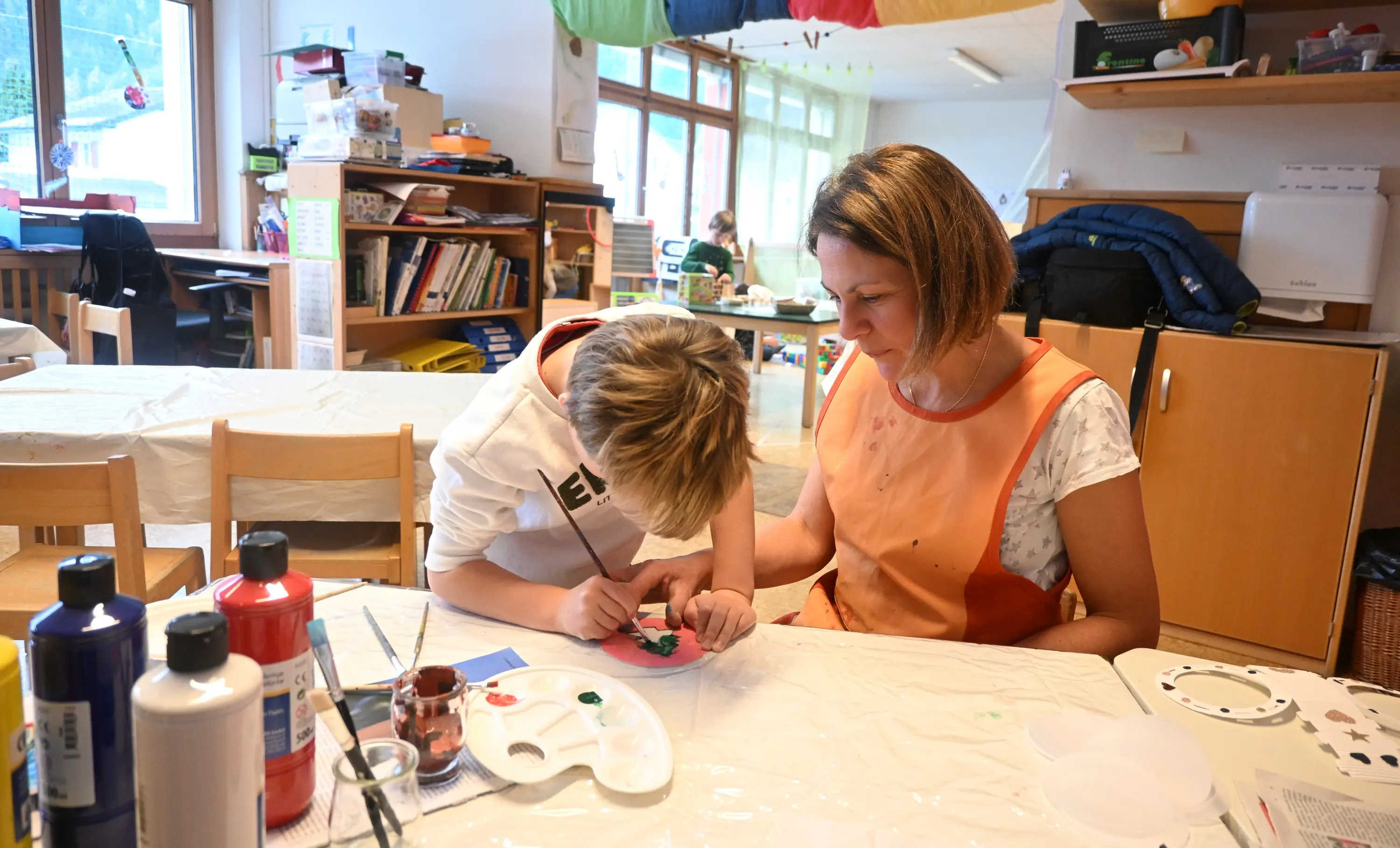 Un bambino sta pitturando con attenzione un oggetto, mentre un adulto lo osserva e lo guida. Entrambi sono seduti a un tavolo in un ambiente luminoso e creativo, cir