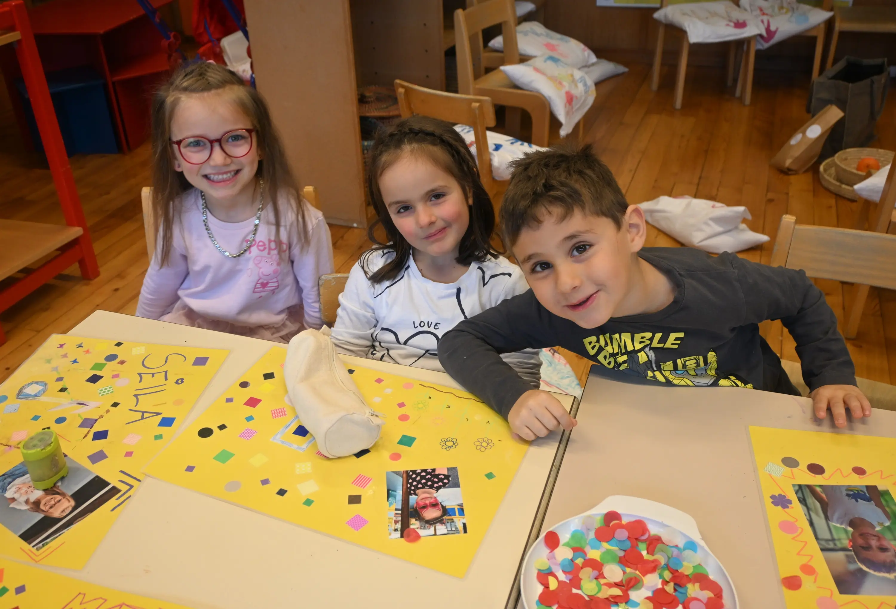 Tre bambini sorridenti seduti a un tavolo, circondati da attività artistiche. Ognuno ha un comportamento vivace mentre lavorano su progetti con materiali colorati come adesivi