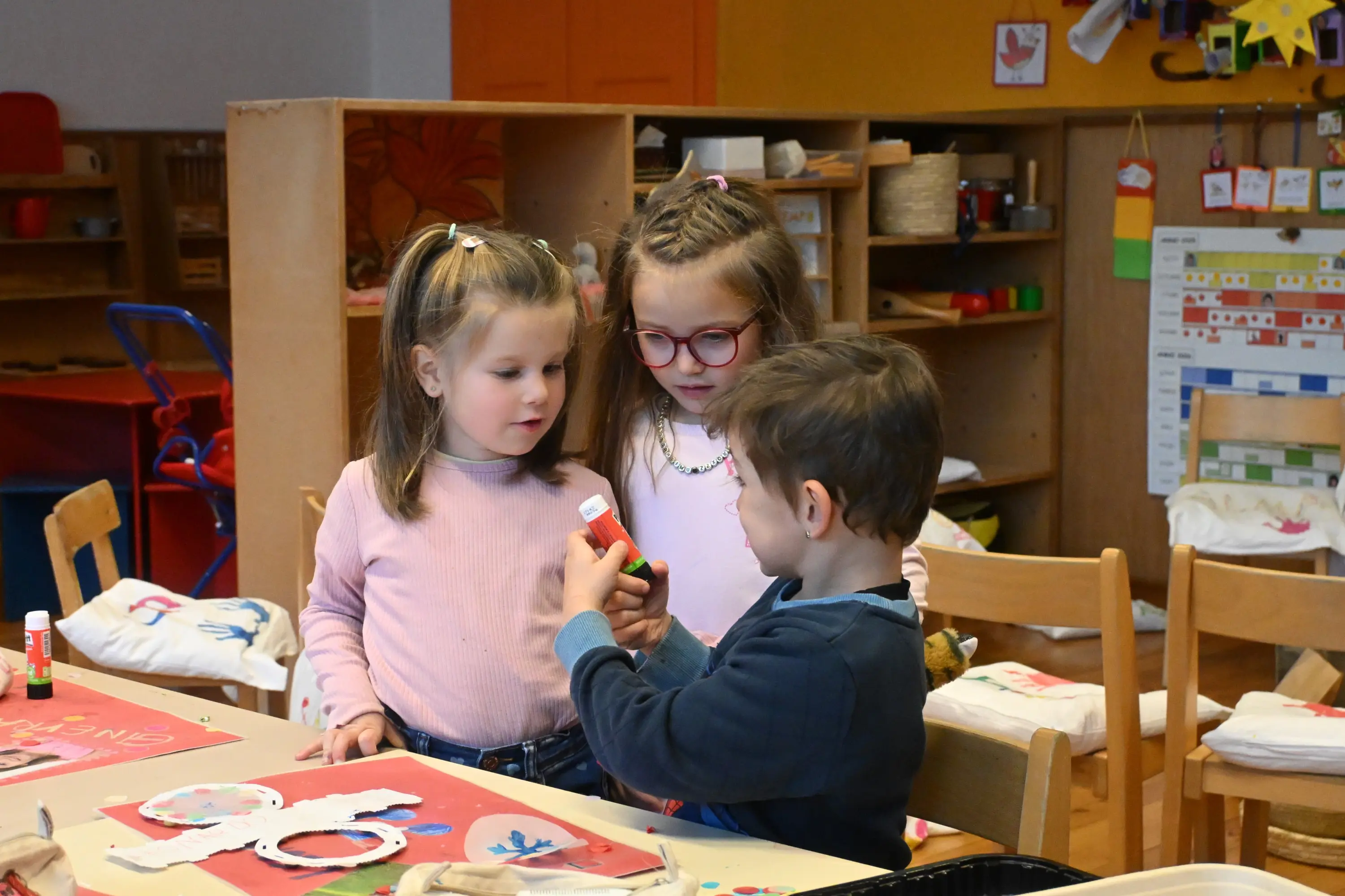 Tre bambini in un’aula colorata stanno condividendo materiali per l’arte. Due ragazze osservano attentamente un ragazzo che tiene una colla. Sul tavolo ci sono pro