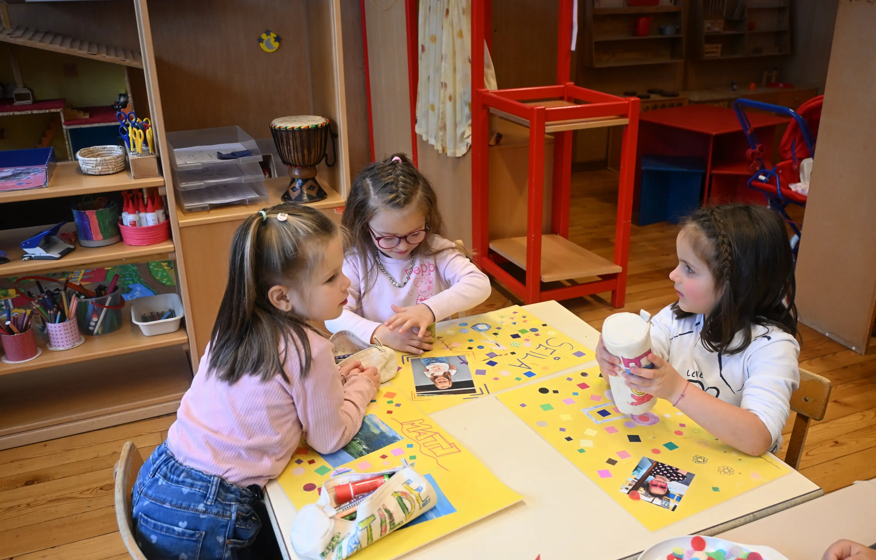 Tre bambine stanno lavorando a un progetto artistico in una stanza per l'infanzia. Sono sedute attorno a un tavolo, utilizzando materiali decorativi come forbici, col