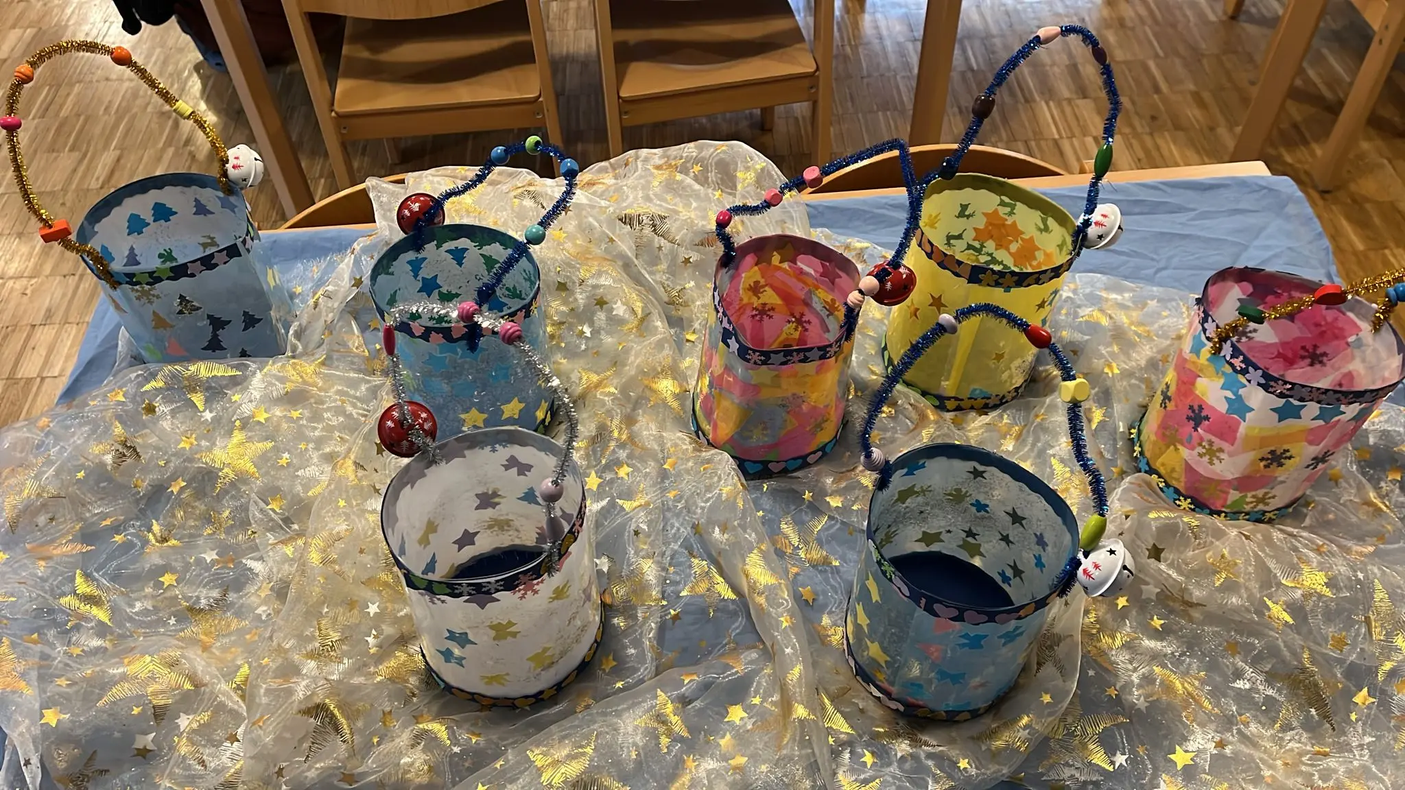 Secchielli decorati con stelle, ciascuno con un manico di perline colorate e campanelli. Sono disposti su un tavolo ricoperto da un tessuto luminos