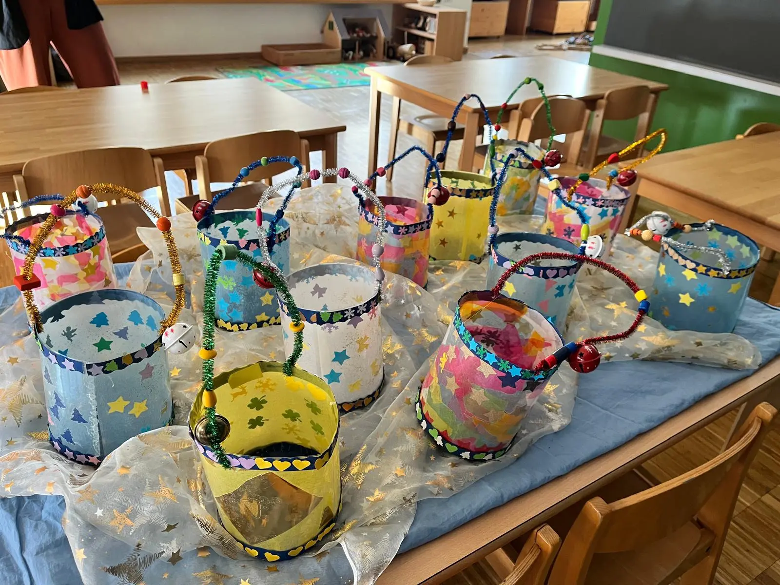 Un tavolo in un ambiente scolastico con vari secchielli decorati in diversi colori, bordati di stelle e con manici di perline. Sono disposti su un blu tessuto