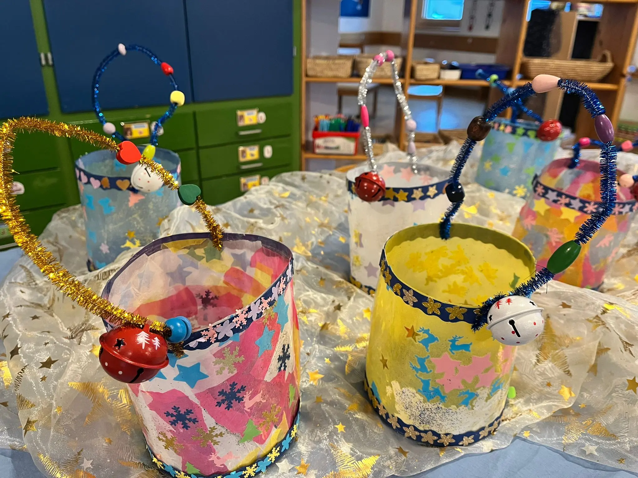 Cinque lanterne colorate decorate con stelle, perline e campanelli, posizionate su una superficie bianca. Sullo sfondo si intravedono scaffali e un ambiente educativo