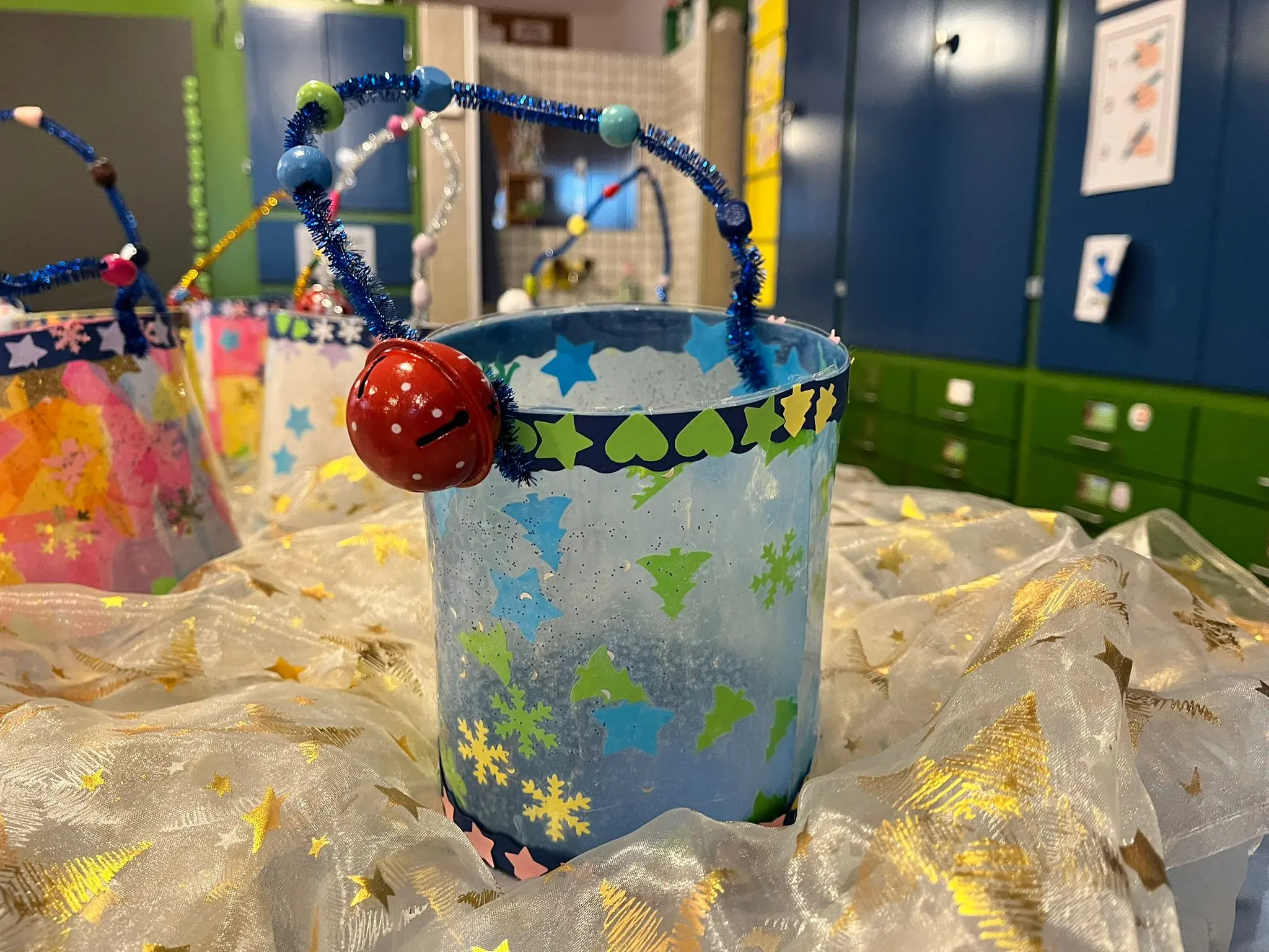 Secchiello decorato con stelle e cuori, ornato da un filo di piPipe colorato e una campanella rossa. Sullo sfondo, altri secchielli simili