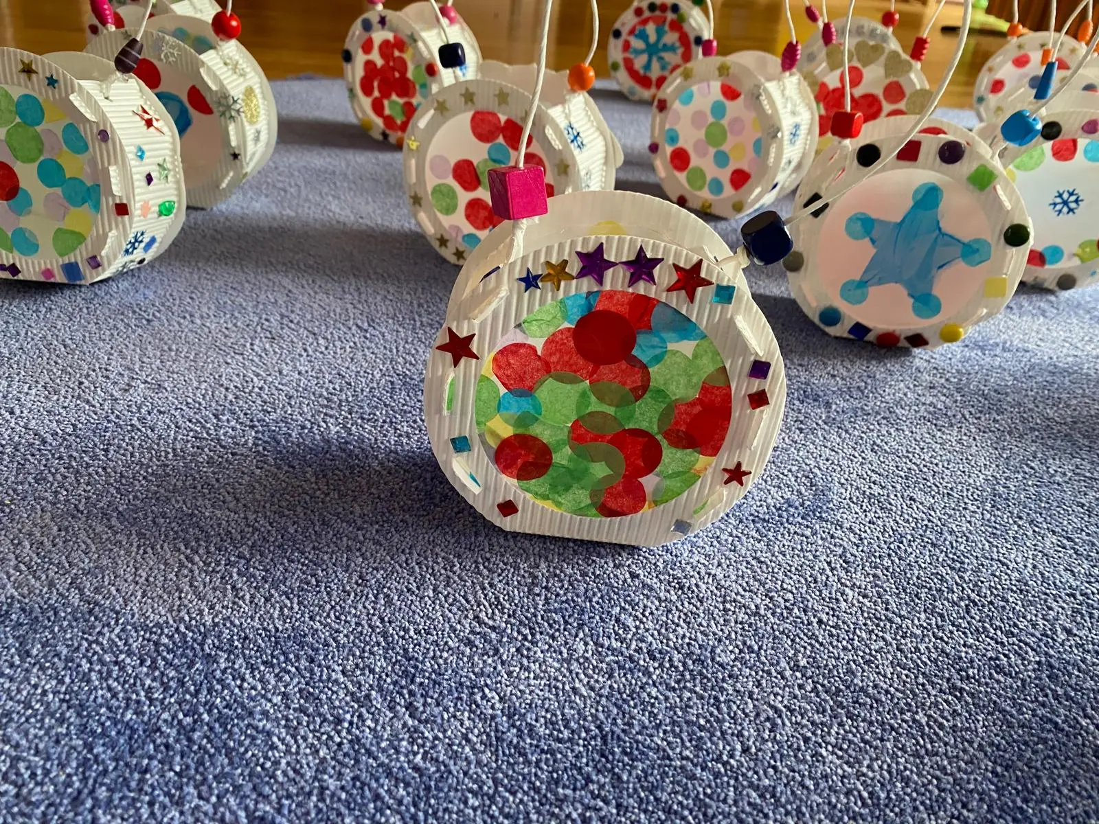 Decorazioni natalizie artigianali a forma di palline, decorate con colori vivaci, puntini e forme. Appese con fili trasparenti su un tappeto blu.