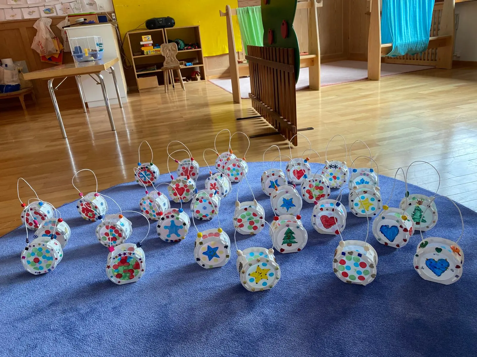 Un gruppo di lanterne artigianali decorate con vari colori e motivi, disposte su un tappeto blu in una stanza per bambini. Sullo sfondo, si intravedono mobili