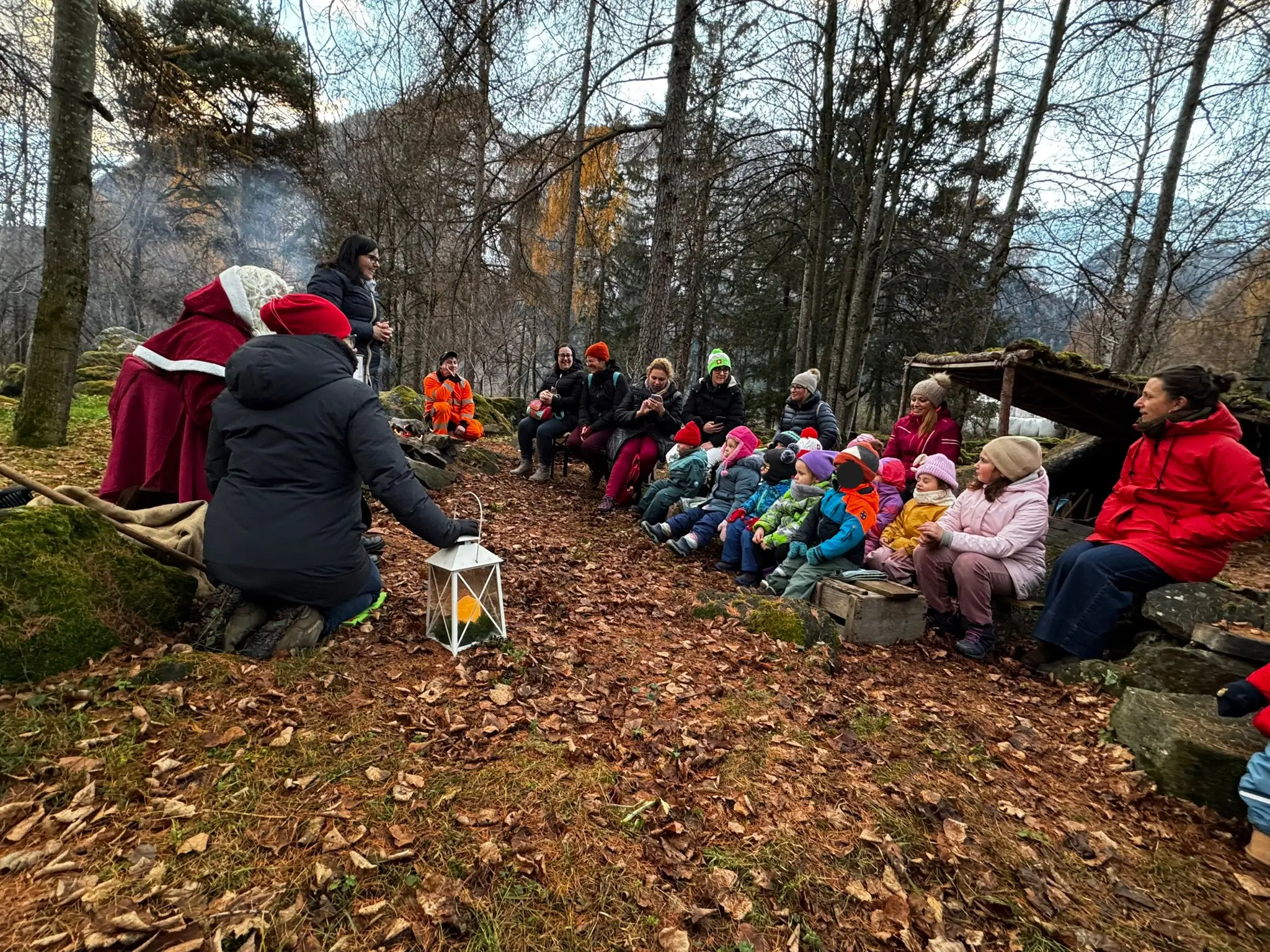 Un gruppo di adulti e bambini si riunisce attorno a un fuoco all'aperto, circondati da alberi. Alcuni indossano abiti invernali e capp