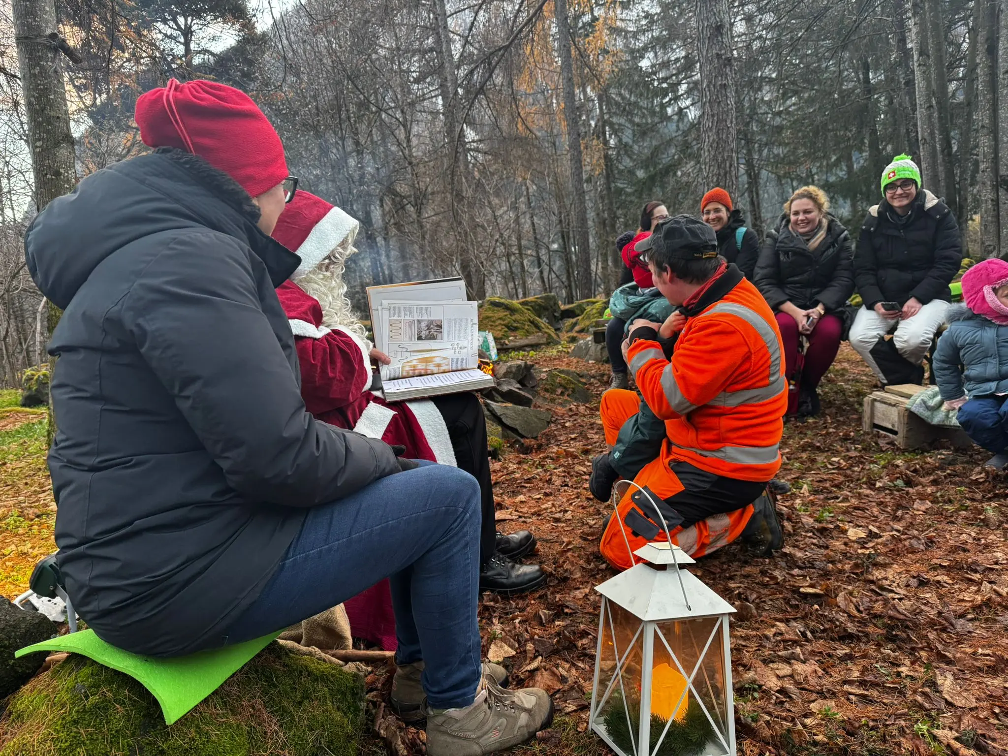 Un gruppo di persone sedute in un bosco autunnale. Una persona in giacca arancione racconta una storia a un piccolo gruppo che ascolta attentamente. Alcuni hanno berre