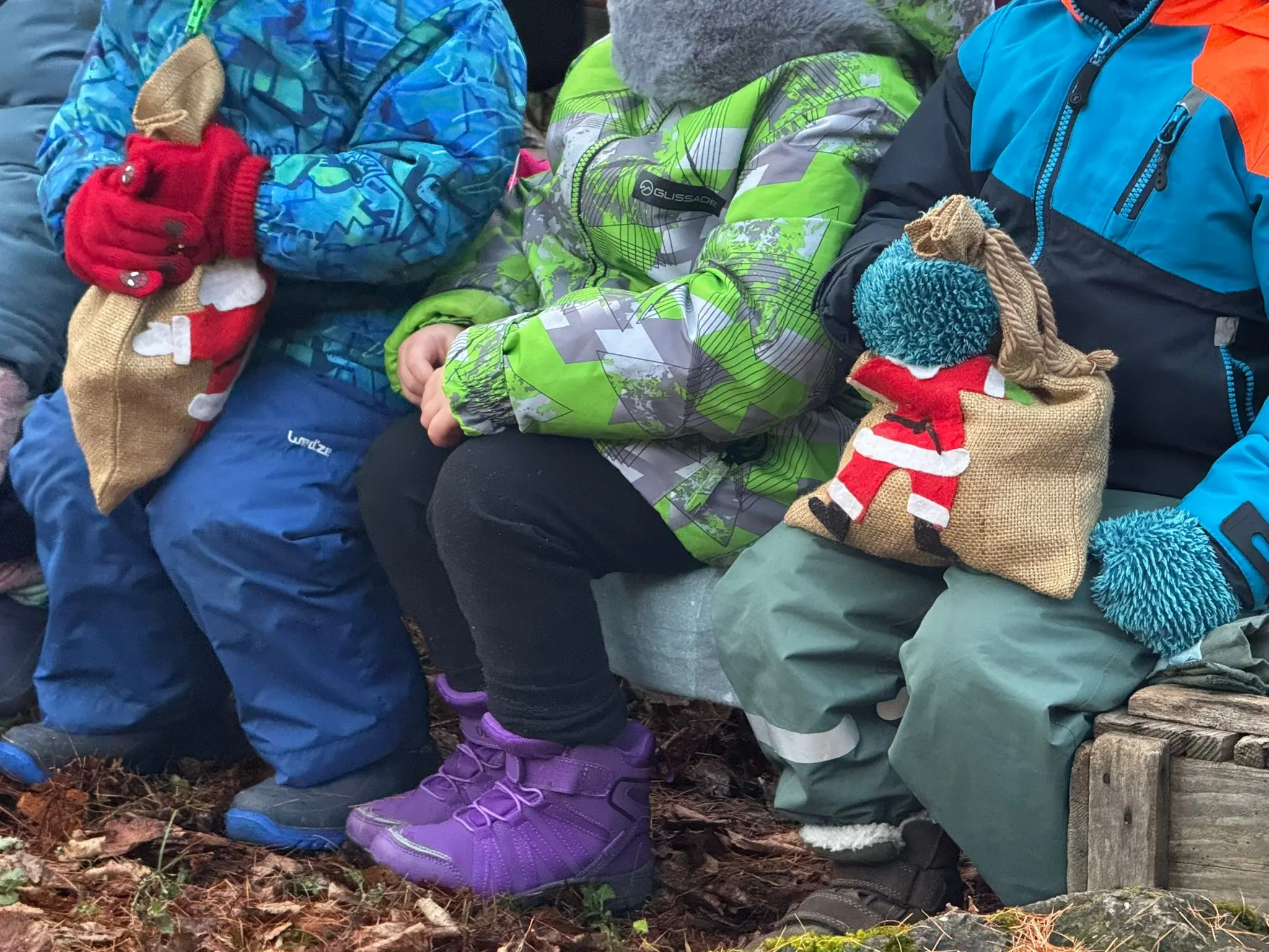 Bambini seduti con sacchetti di stoffa in mano, indossano giacche colorate e guanti. Il sacchetto di uno dei bambini presenta un disegno di