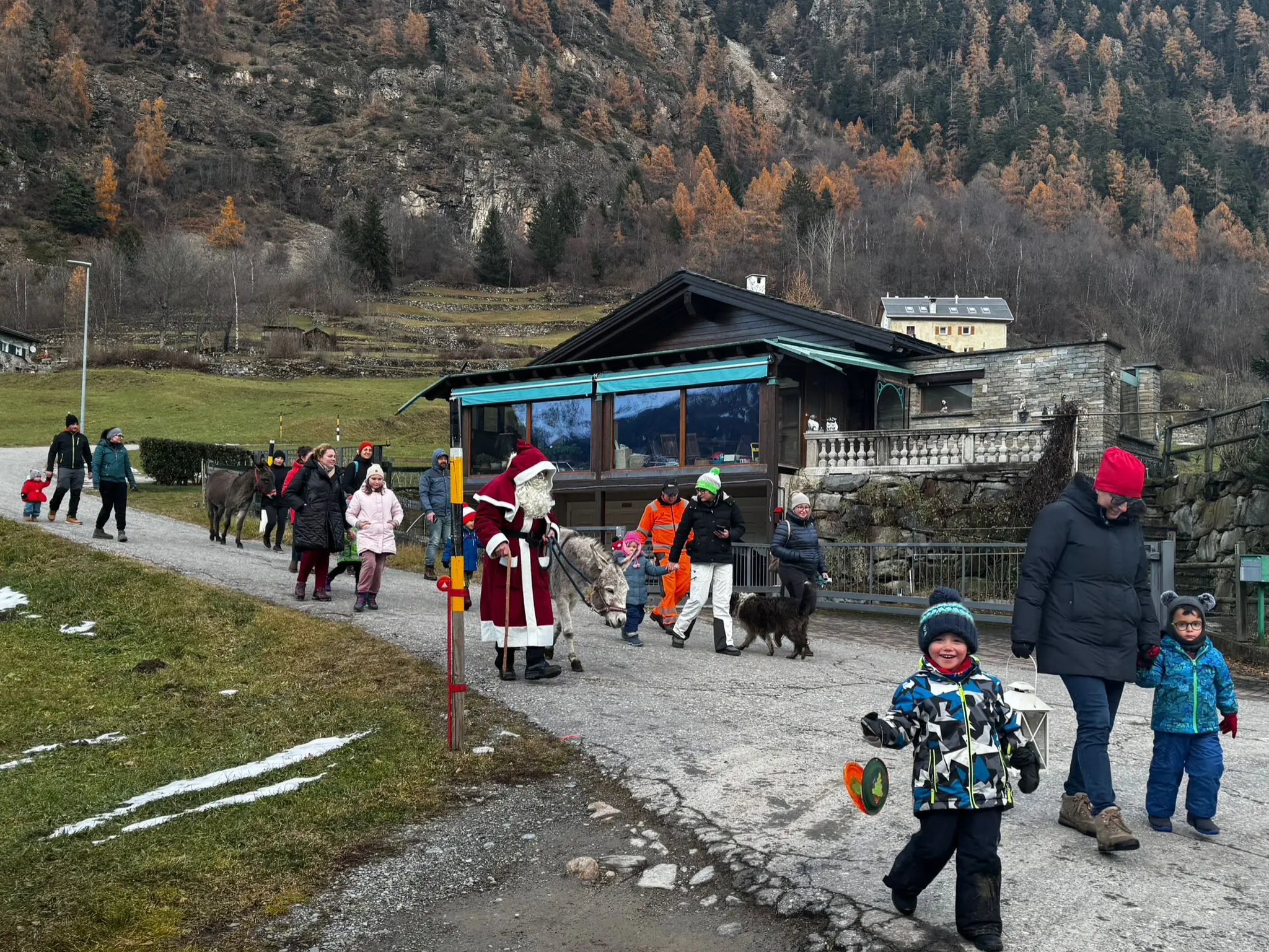 Un gruppo di persone che cammina lungo una strada, tra cui un uomo vestito da Babbo Natale, contornati da animali e un ambiente montano. Alcuni bambini sorridono
