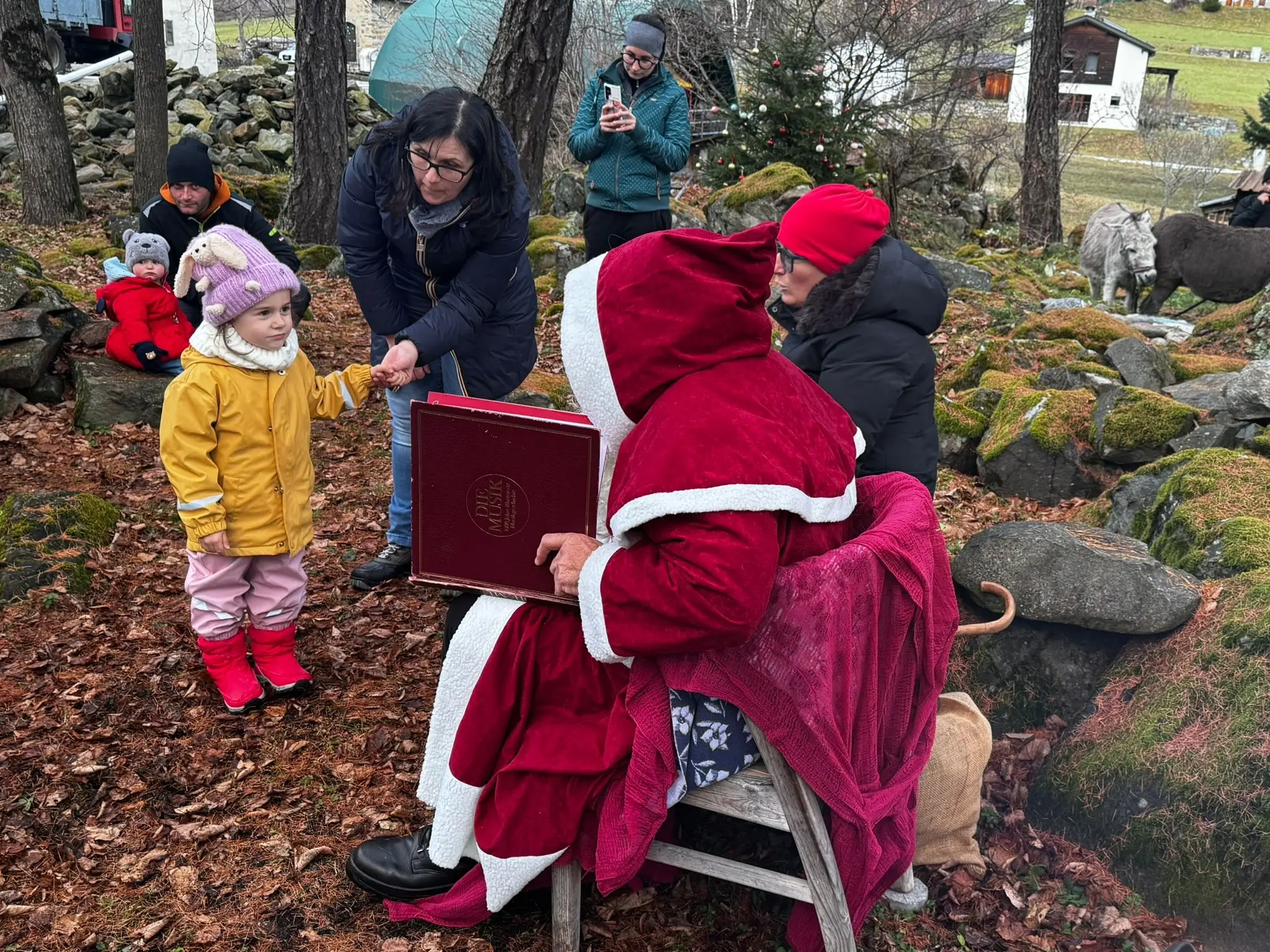 Una persona vestita da Babbo Natale seduta su una sedia, mentre una bambina in giacca gialla e stivali rossi si avvicina per ricevere un regalo. Al