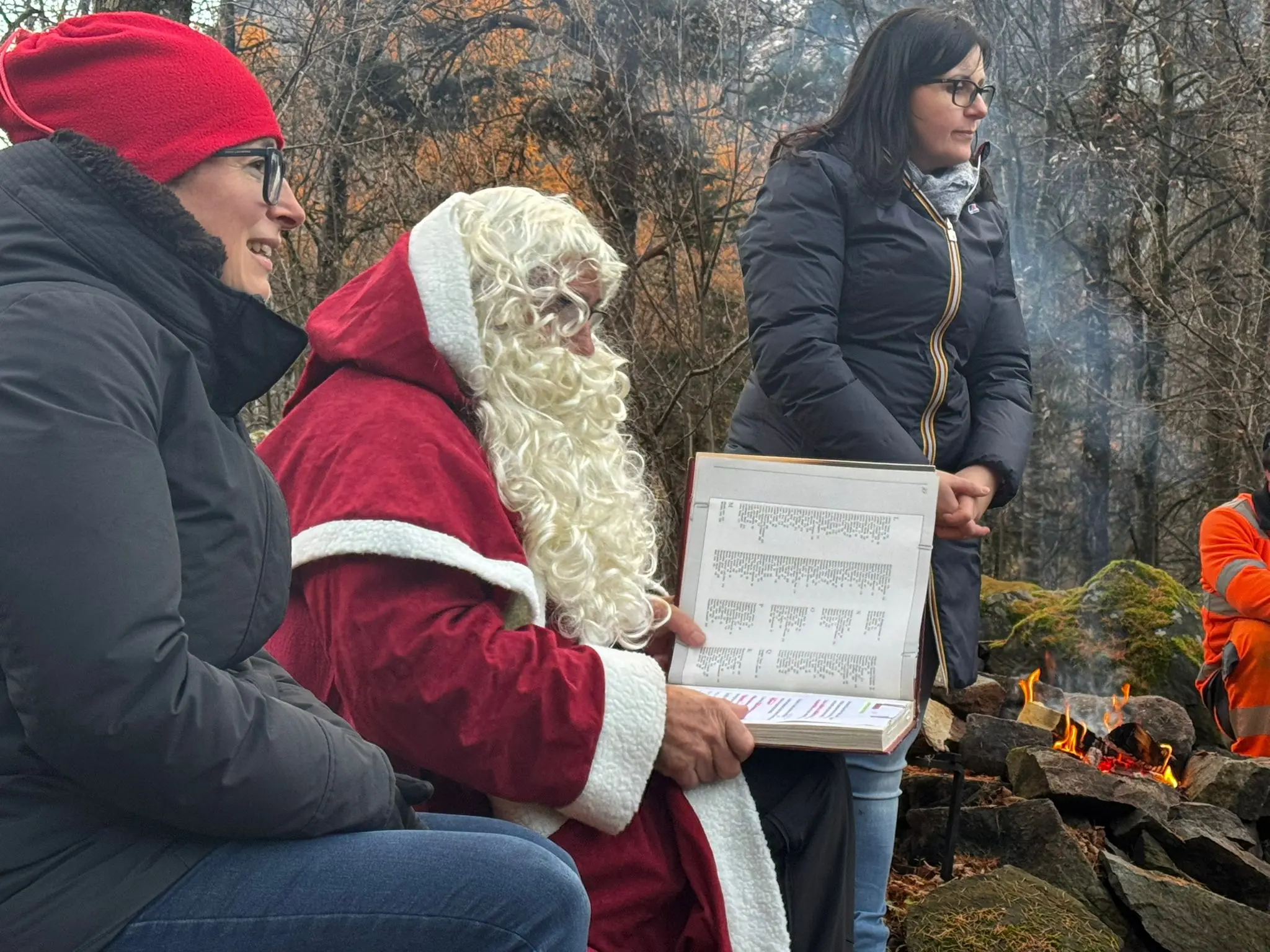 Due persone sorridenti sedute accanto a un uomo vestito da Babbo Natale, che tiene un libro aperto. Sullo sfondo, un fuoco acceso e alberi.