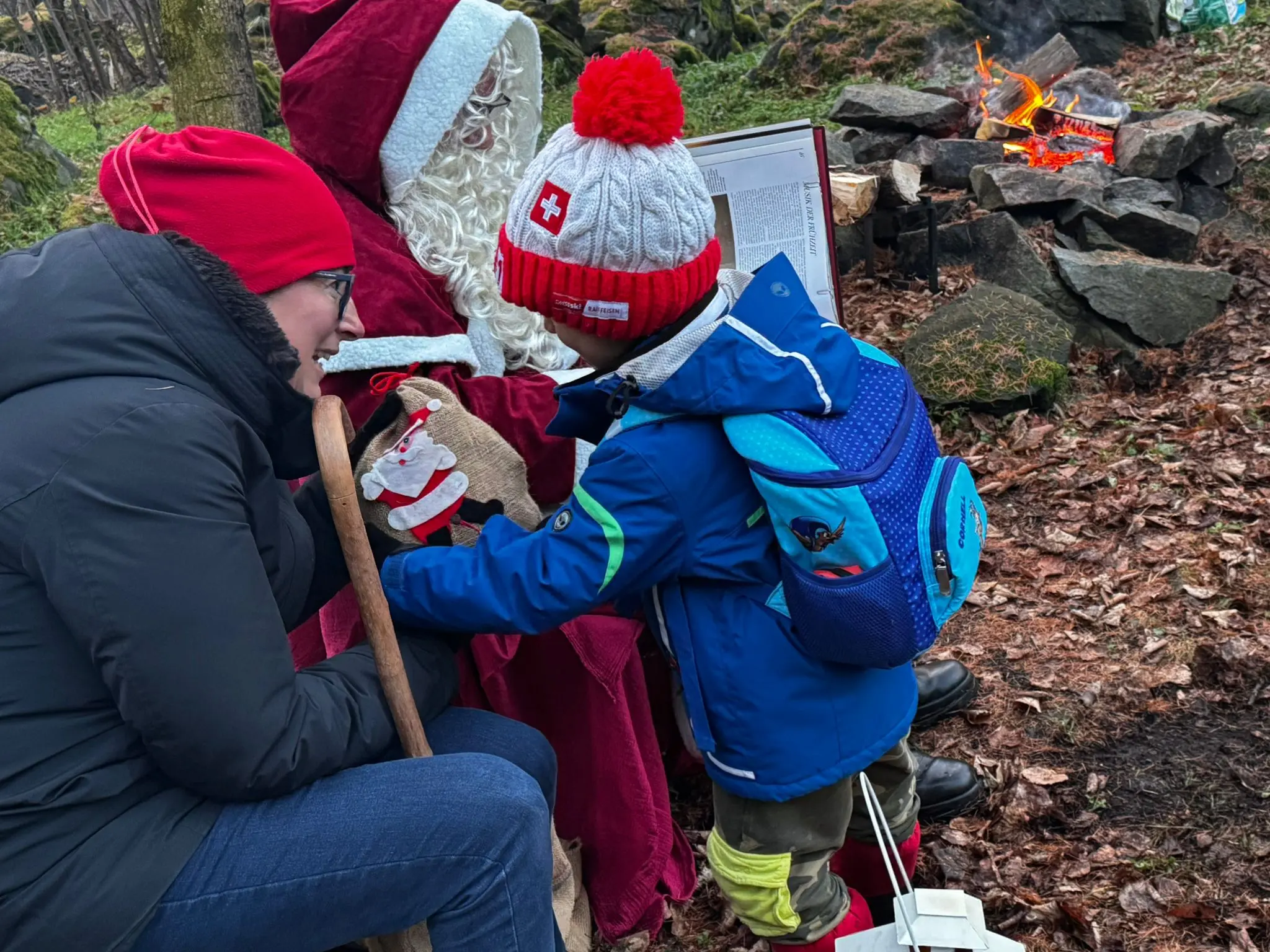 Una mamma e un bambino incontrano Babbo Natale nel bosco, vicino a un falò. Il bambino, con uno zaino blu, tocca il sacco di Babbo Natale mentre entr