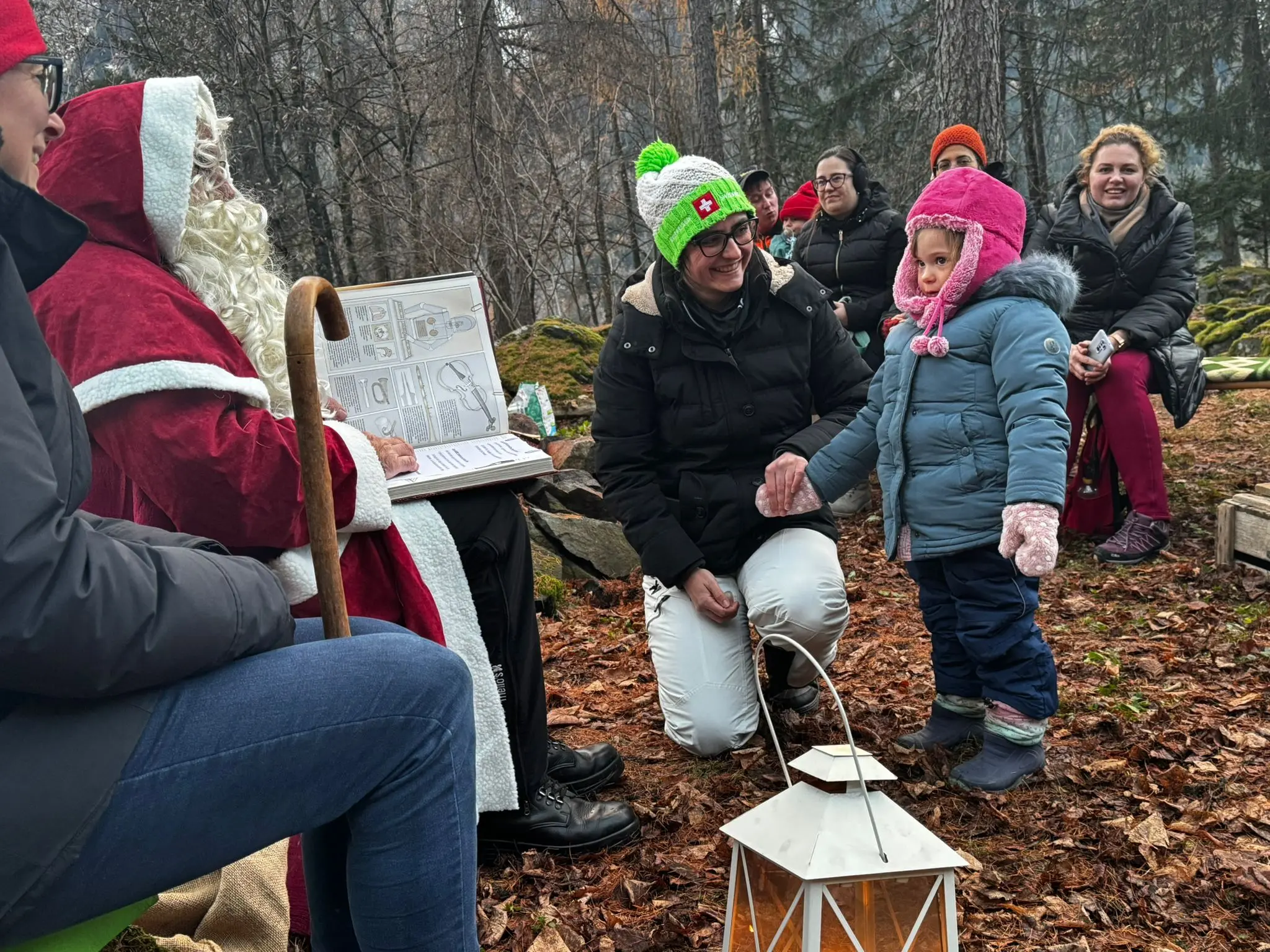 Babbo Natale seduto su una panchina nel bosco, circondato da adulti e bambini. Una bambina con un cappotto blu e un cappello rosa si avvicina