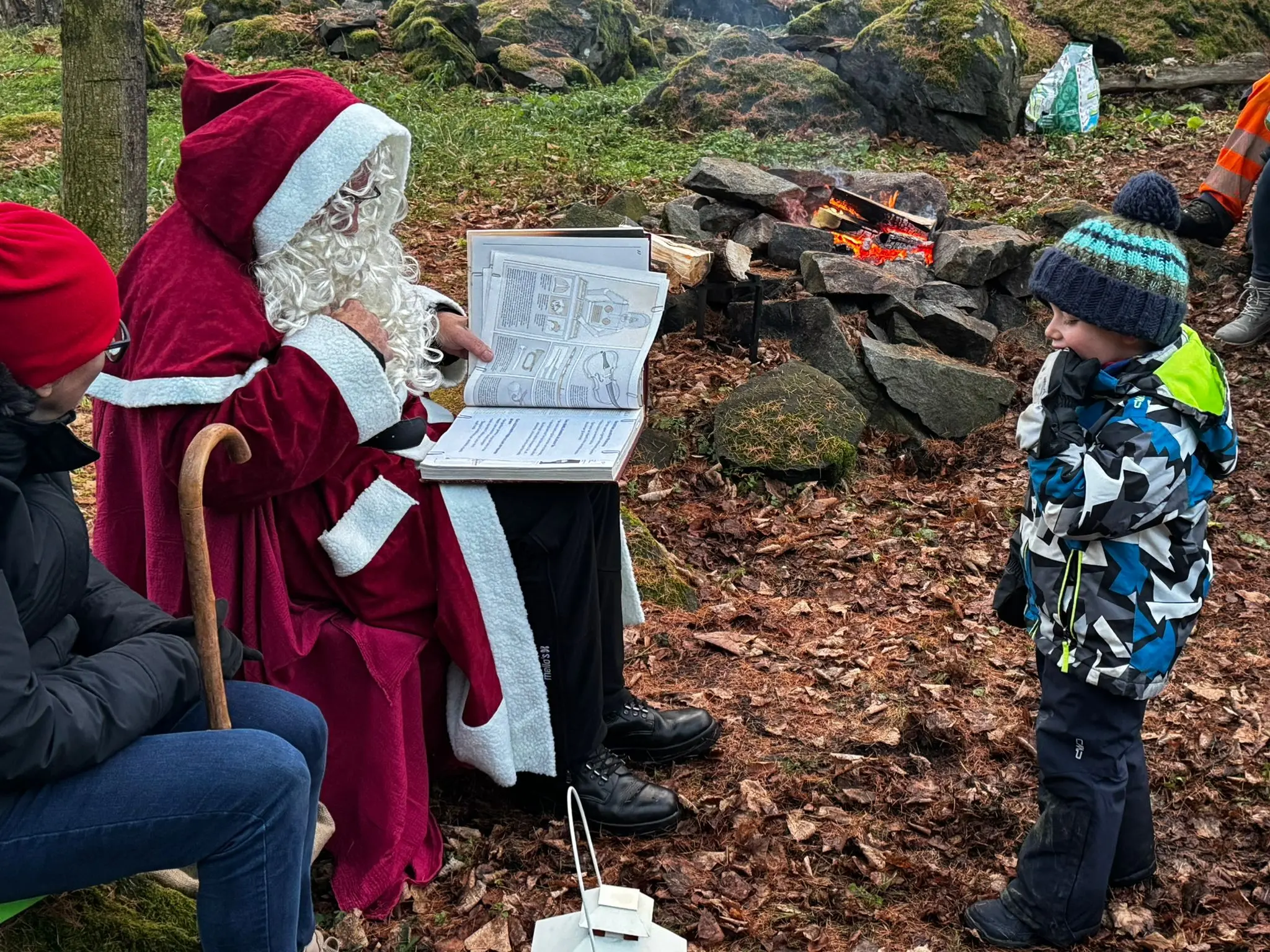 Babbo Natale legge un libro a un bambino vestito con giacca a colori vivaci, mentre una donna ascolta seduta accanto. Un falò scoppietta tra le rocce circ