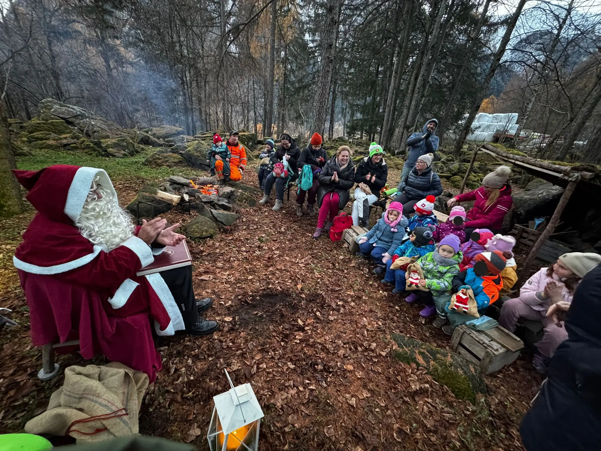 Babbo Natale racconta storie attorno a un fuoco, circondato da bambini e adulti seduti su un terreno boschivo. Gli alberi e la vegetazione autunn
