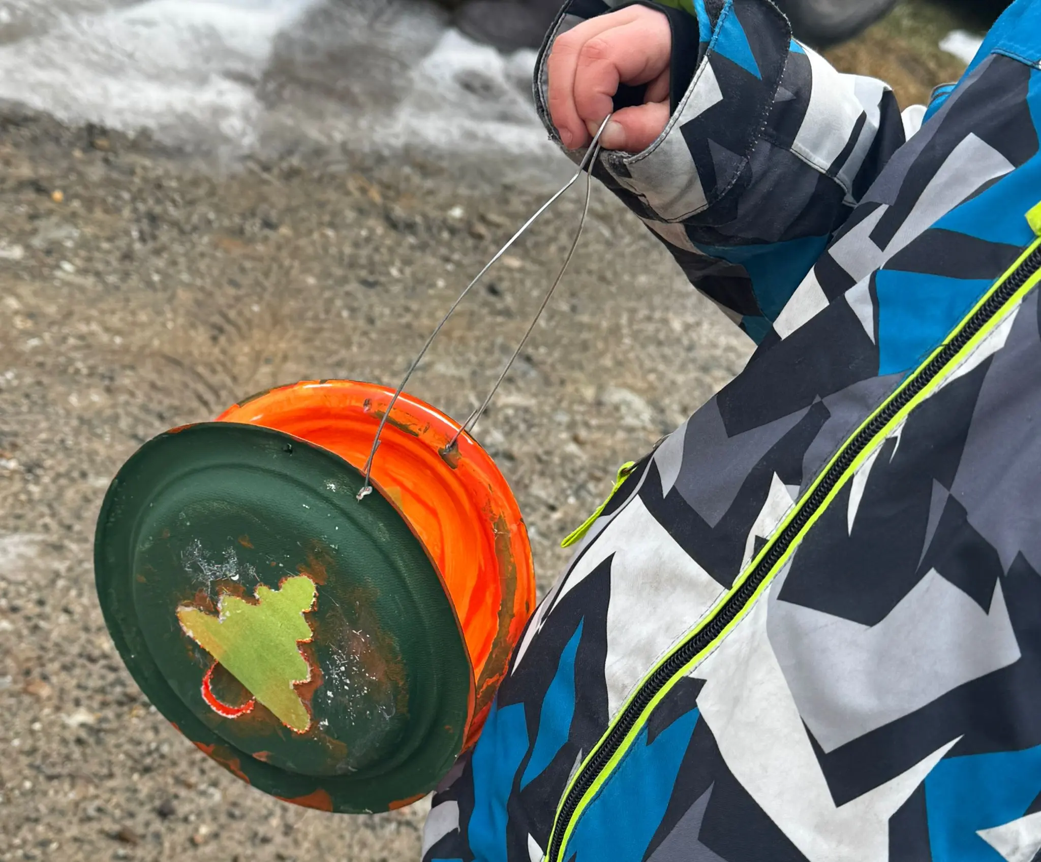 Un bambino tiene in mano un contenitore di plastica arancione e verde, decorato con un albero di Natale dorato. Indossa un giubbotto invernale con un