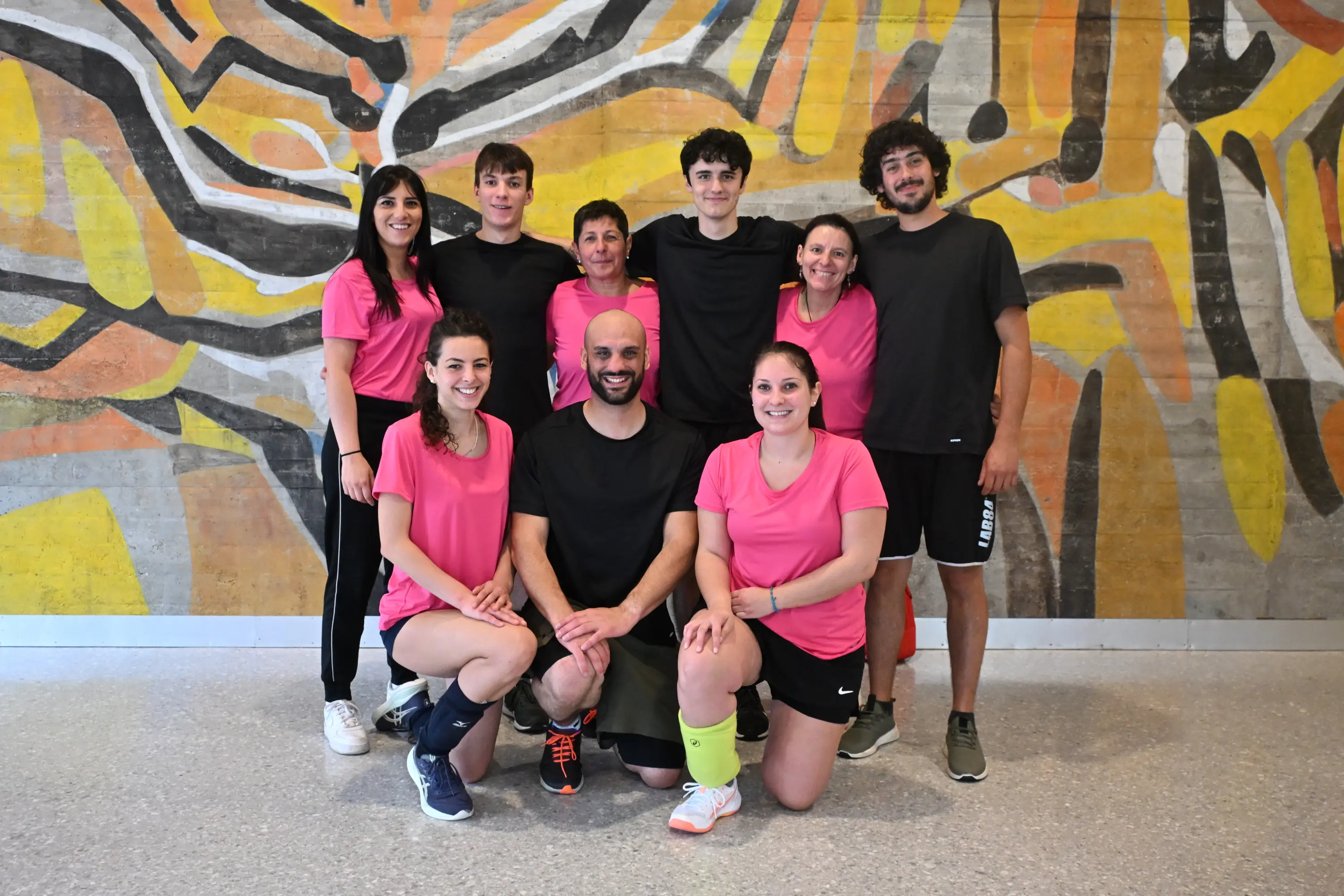 Gruppo di otto persone sorridenti che indossano magliette rosa, con un uomo al centro. Sono posizionati davanti a un muro decorato con un murale colorato.