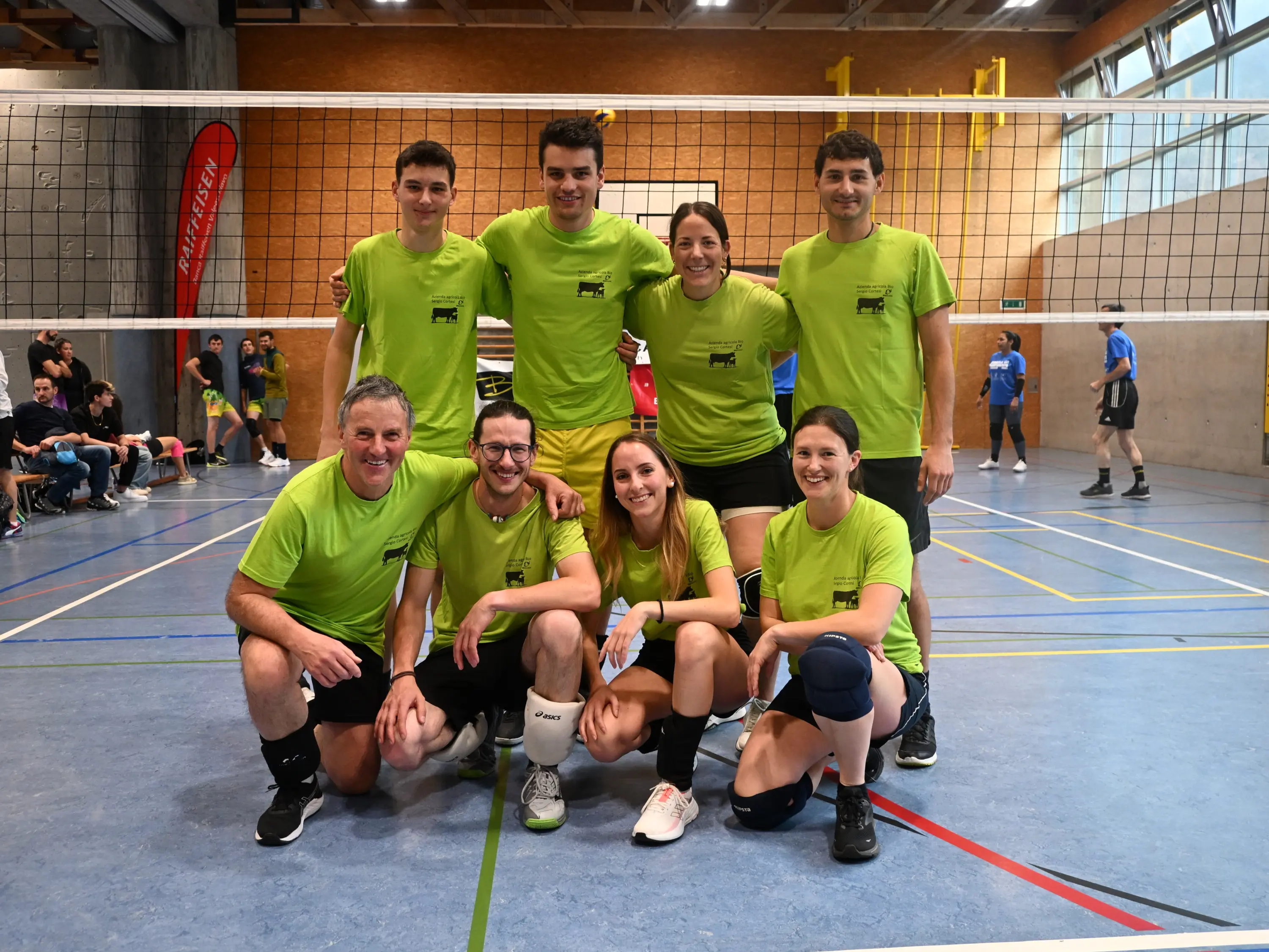 Un gruppo di otto giocatori di volley in magliette verdi, sorridenti, posano insieme in palestra. Sullo sfondo si vede una rete da volley e alcune persone che giocano.