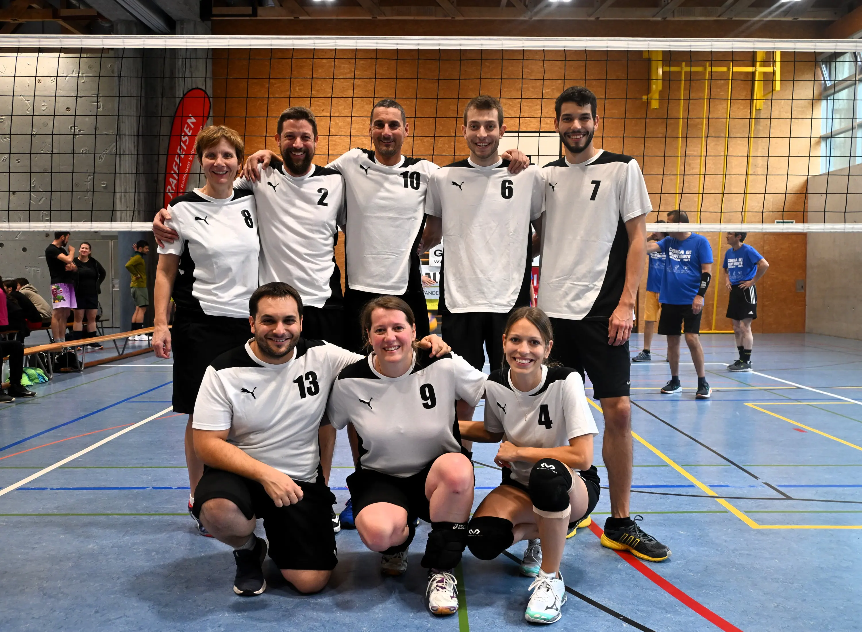 Squadra di pallavolo composta da otto membri, sorridenti, in una palestra. Sono vestiti con magliette bianche e pantaloni neri, alcuni hanno numeri sulla maglietta. Sullo sfondo si vede una rete da pallavolo.