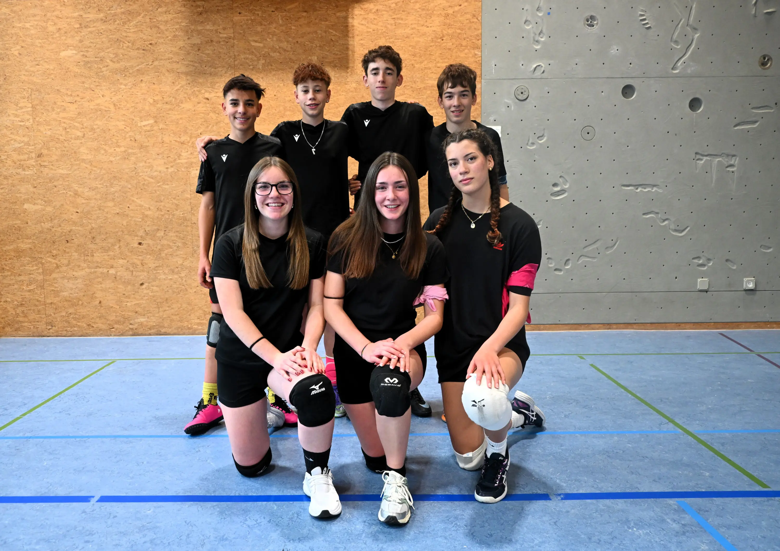 Un gruppo di sei giovani atleti in abbigliamento sportivo nero, sorridenti e in posa in una palestra. Due ragazze sono in primo piano, inginocchiate, mentre i ragazzi sono in piedi sullo sfondo. Tutti indossano calzini e ginoc