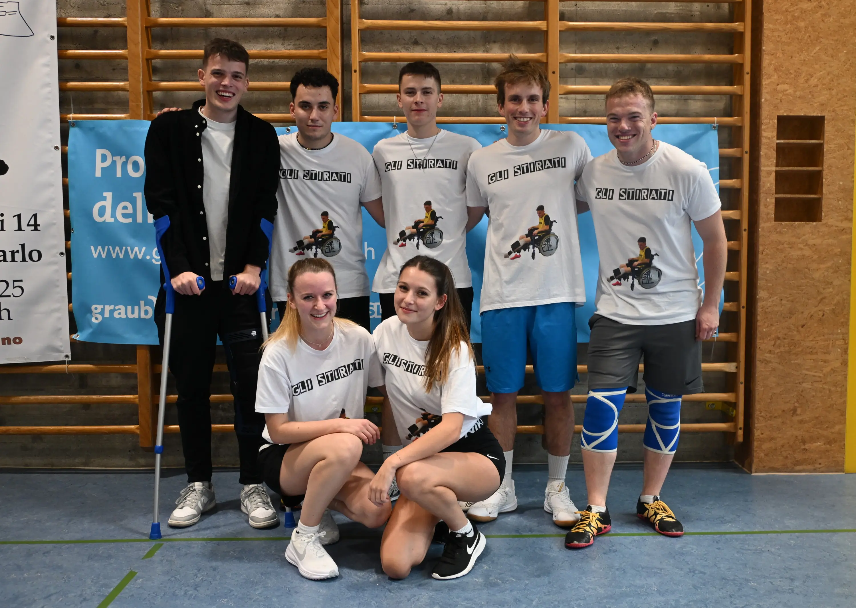 Un gruppo di sei ragazzi sorridenti, vestiti con magliette bianche con la scritta "2U SIRPANI", si trova su un campo da palestra. Due di loro sono in piedi, mentre quattro sono in ginocchio; uno di loro tiene delle stampelle.