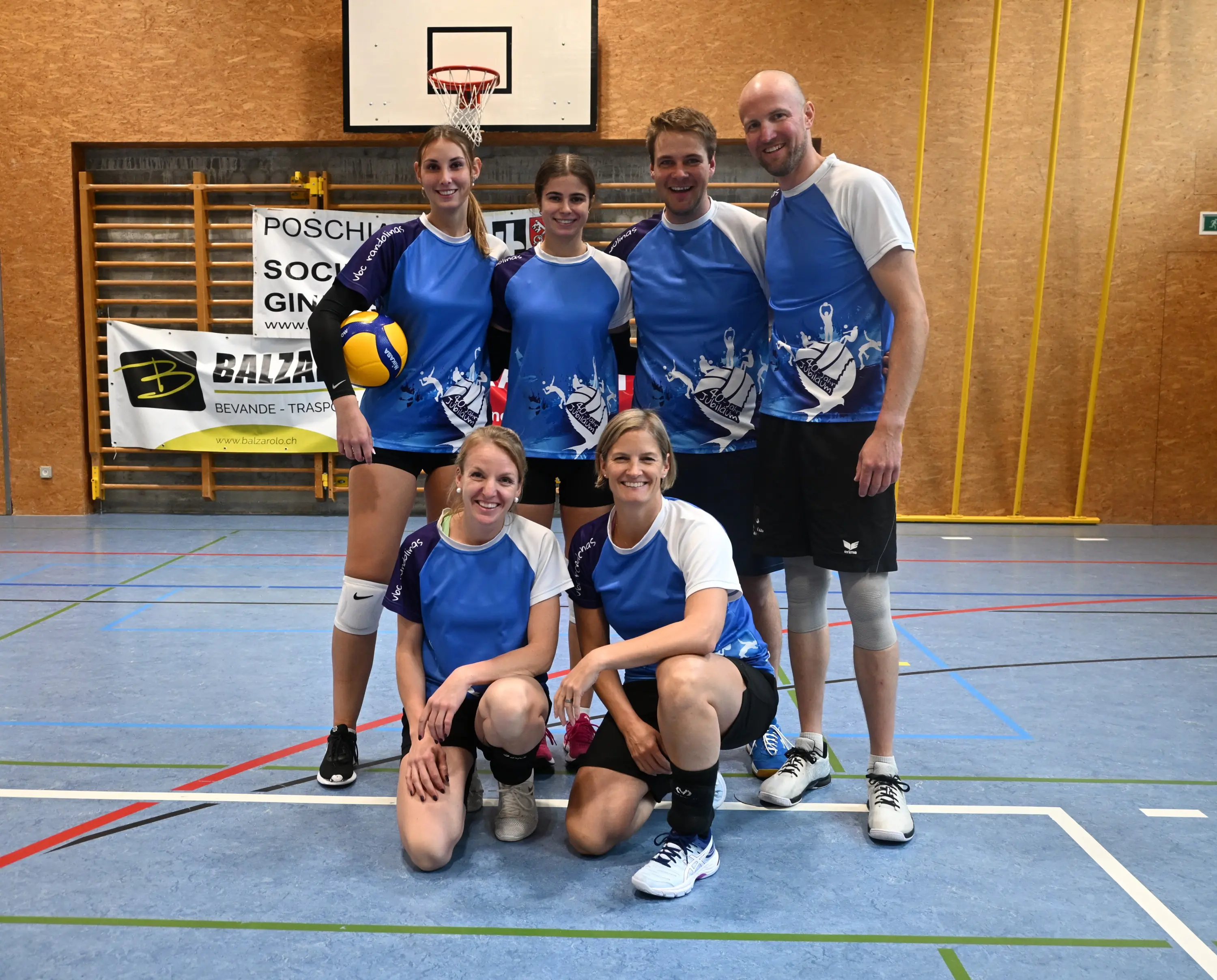 Un gruppo di sei giocatori di pallavolo in maglietta blu e bianca, sorridenti, posano in un palazzetto dello sport. Sullo sfondo si vede un canestro e una bandiera sponsor.