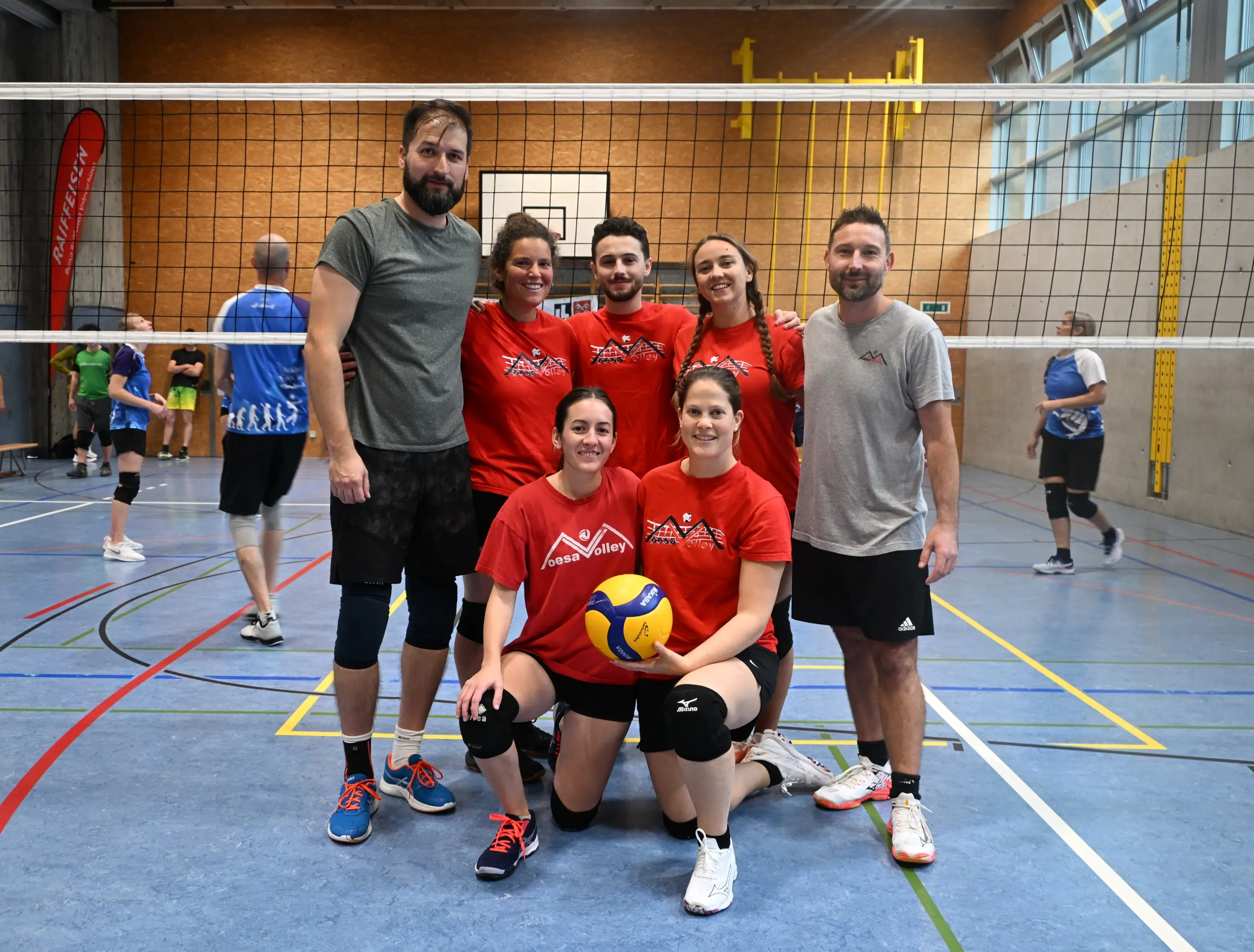 Un gruppo di sette persone con magliette rosse e pantaloni sportivi, posano insieme in un campo di pallavolo. Alcuni membri sono in piedi, mentre altri sono accovacciati in prima fila. Sullo sfondo, si vedono altri giocatori in azione