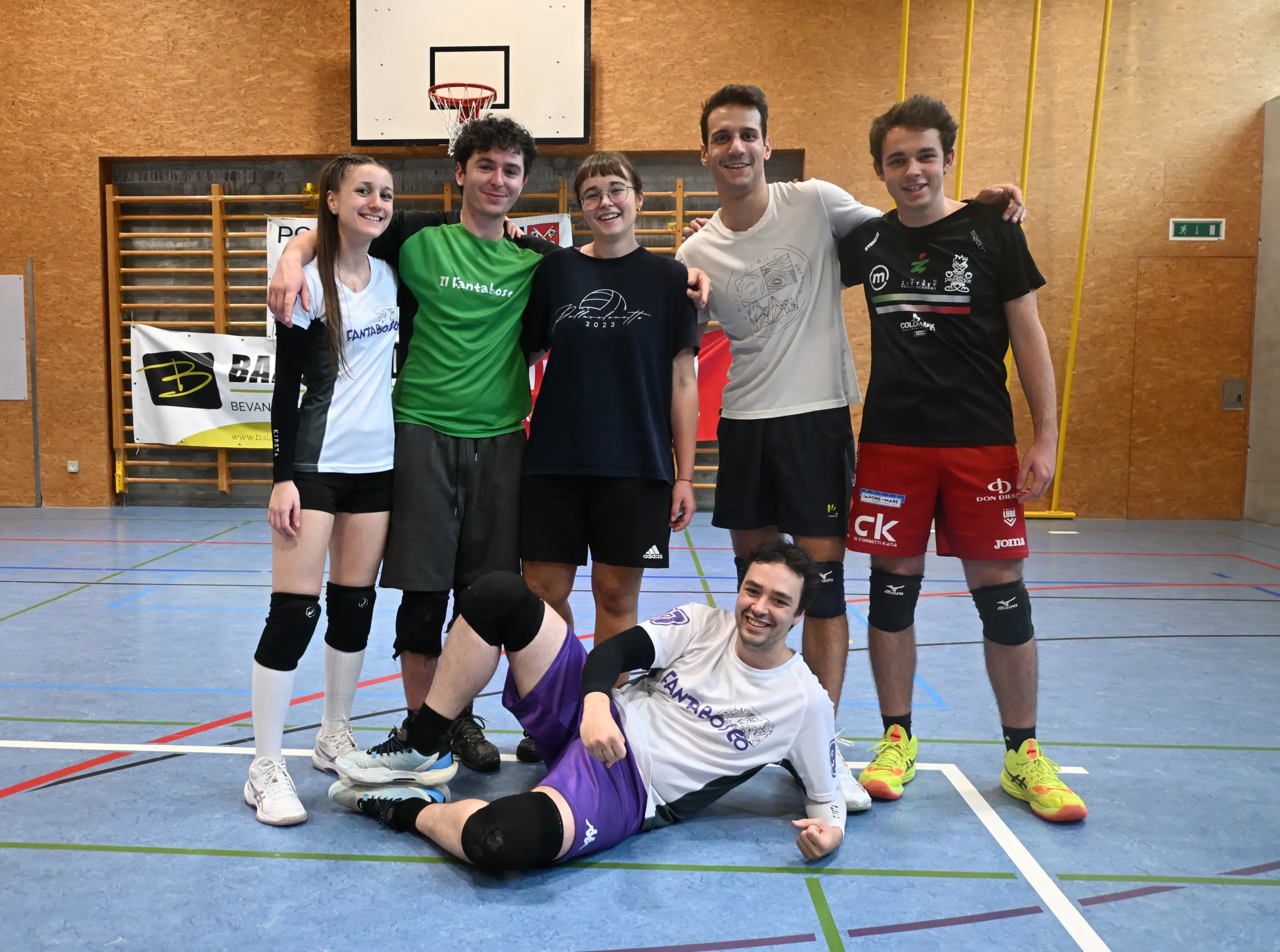 Un gruppo di sei giovani giocatori di pallavolo si trova in un campo sportivo. Cinque stanno in piedi, sorridendo e abbracciandosi, mentre uno è accovacciato sul pavimento. Indossano maglie e pantaloni sportivi di diversi colori.