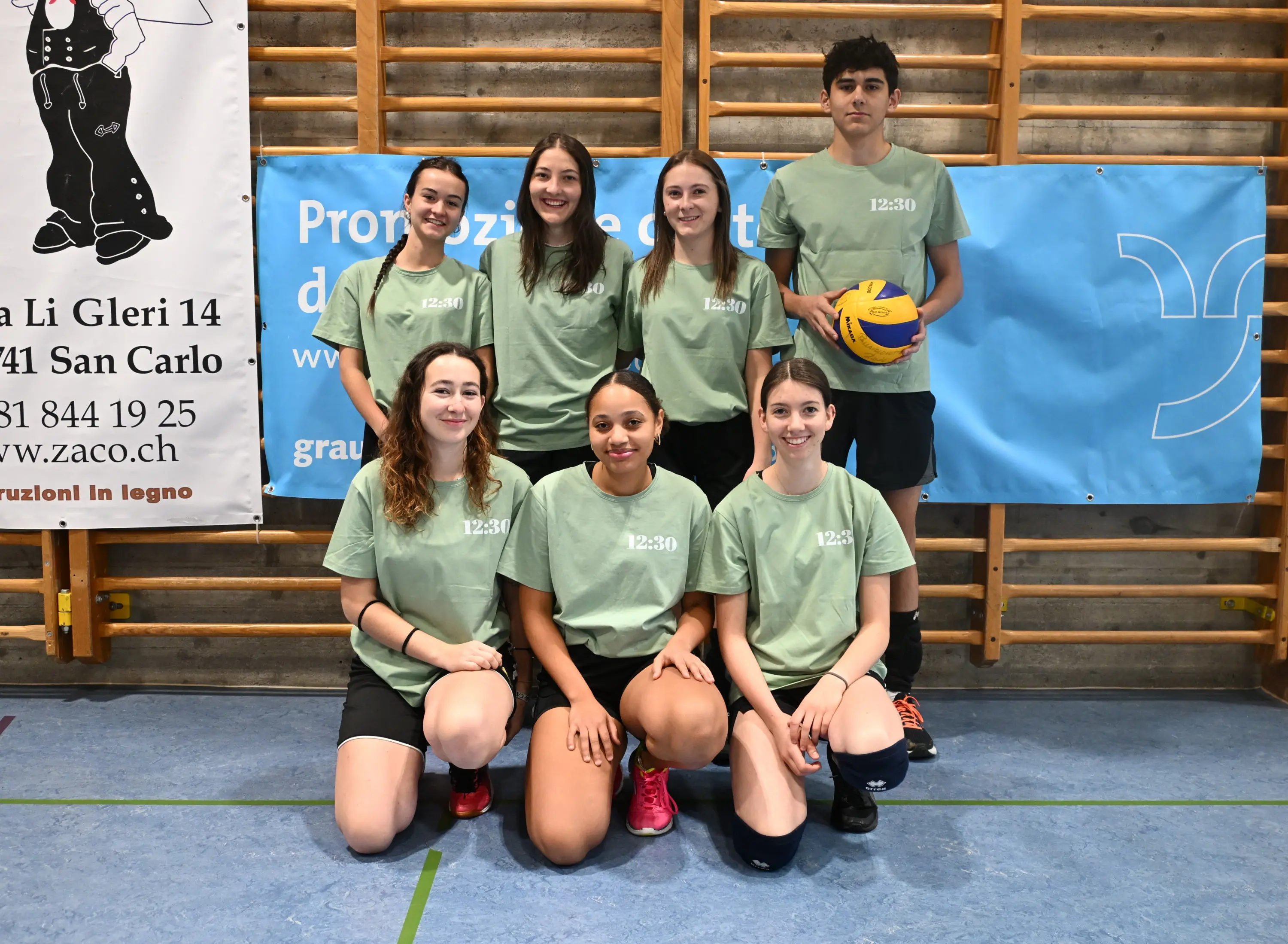 Un gruppo di sette giovani atleti, tutte donne tranne un uomo, indossa magliette verdi e pantaloni corti neri. Sono in piedi in una palestra con un pallone da pallavolo. Sullo sfondo ci sono striscioni e una parete di legno