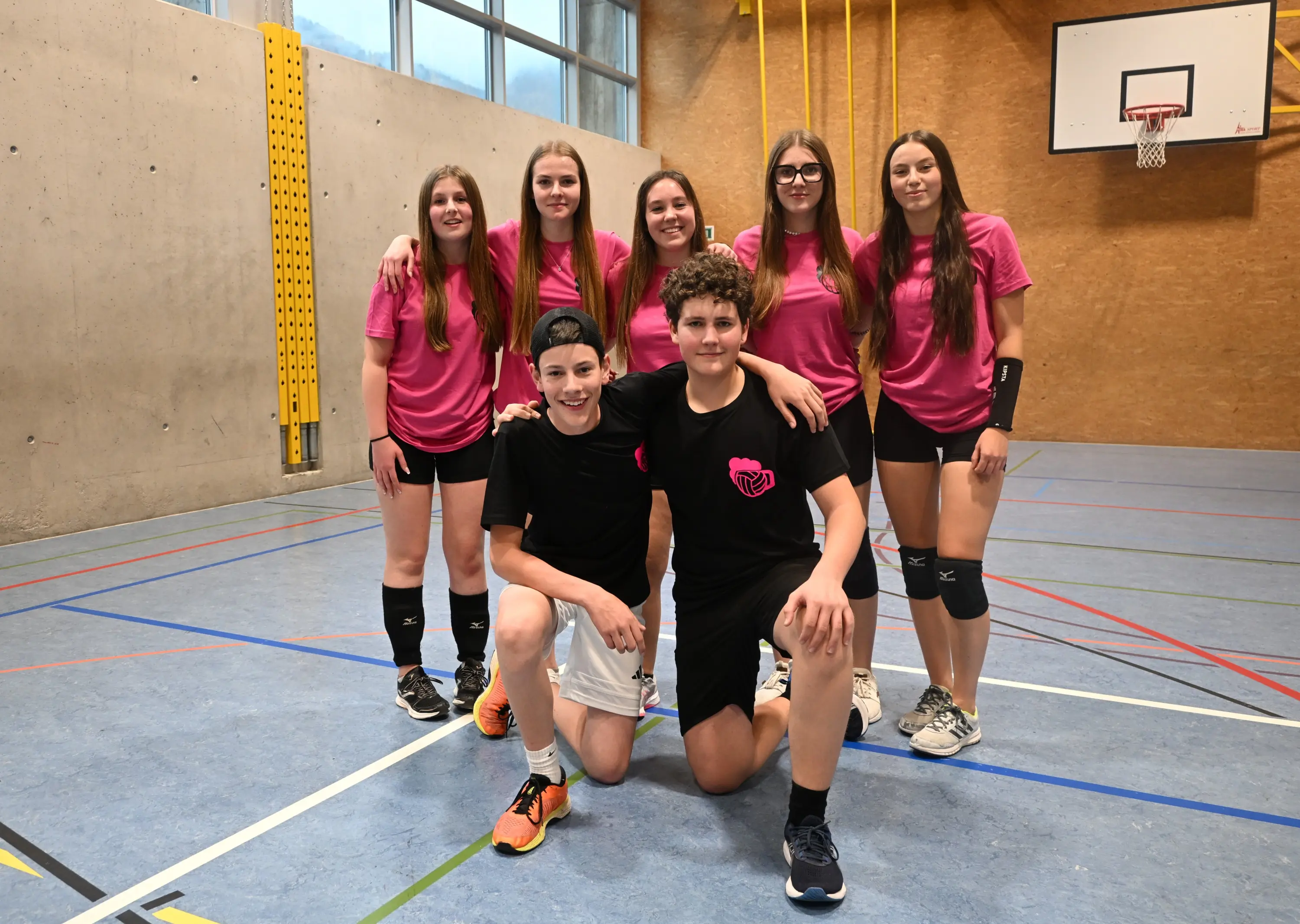 Un gruppo di sei ragazzi e ragazze in abbigliamento sportivo rosanero posano insieme in un campo di pallavolo. Due ragazzi sono in primo piano, uno in ginocchio e l'altro in piedi al centro, mentre le ragazze si trovano dietro di