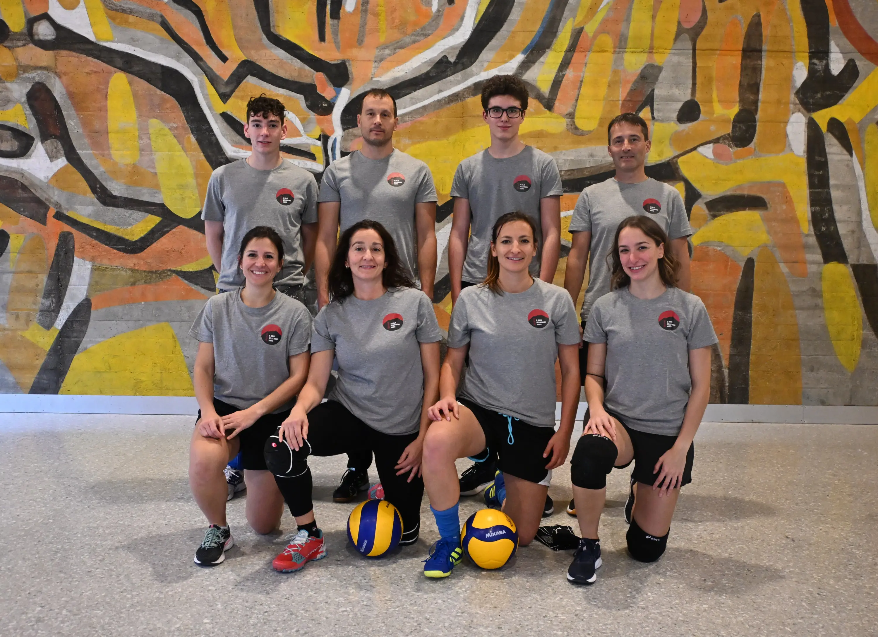 Un gruppo di otto persone, in piedi e inginocchiate, indossa magliette grigie con un logo e pantaloni sportivi. Alcuni stanno tenendo un pallone da pallavolo. Lo sfondo presenta un murale colorato con tonalità gialle