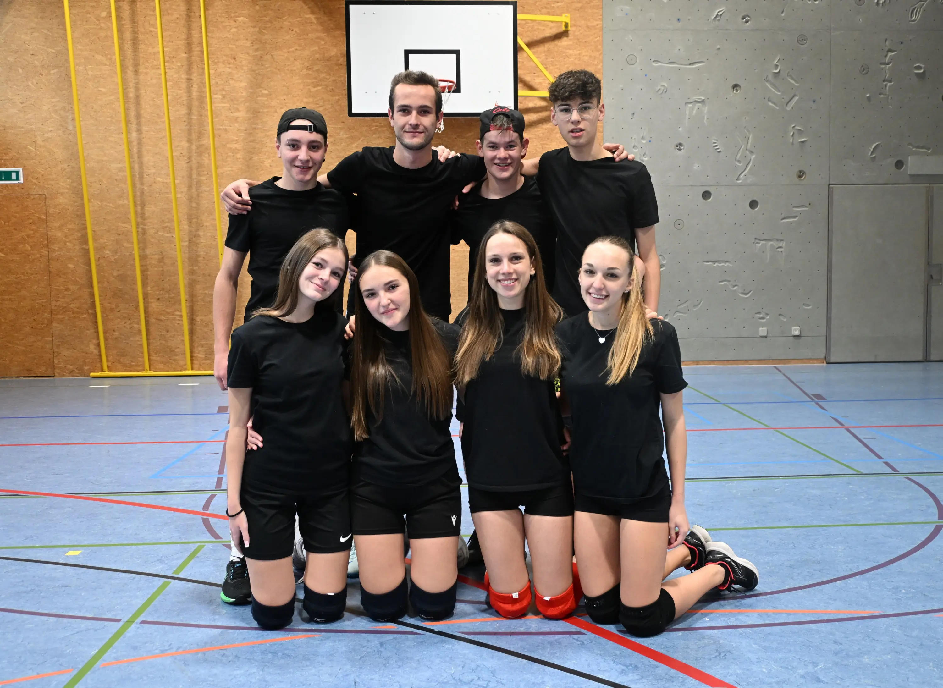 Un gruppo di nove giovani atleti in maglietta nera e pantaloni sportivi si trova in una palestra. Sono in posizione di squadra, con alcuni in piedi e altri accovacciati. Il pavimento è blu e ci sono elementi di pallavolo sullo sfondo.