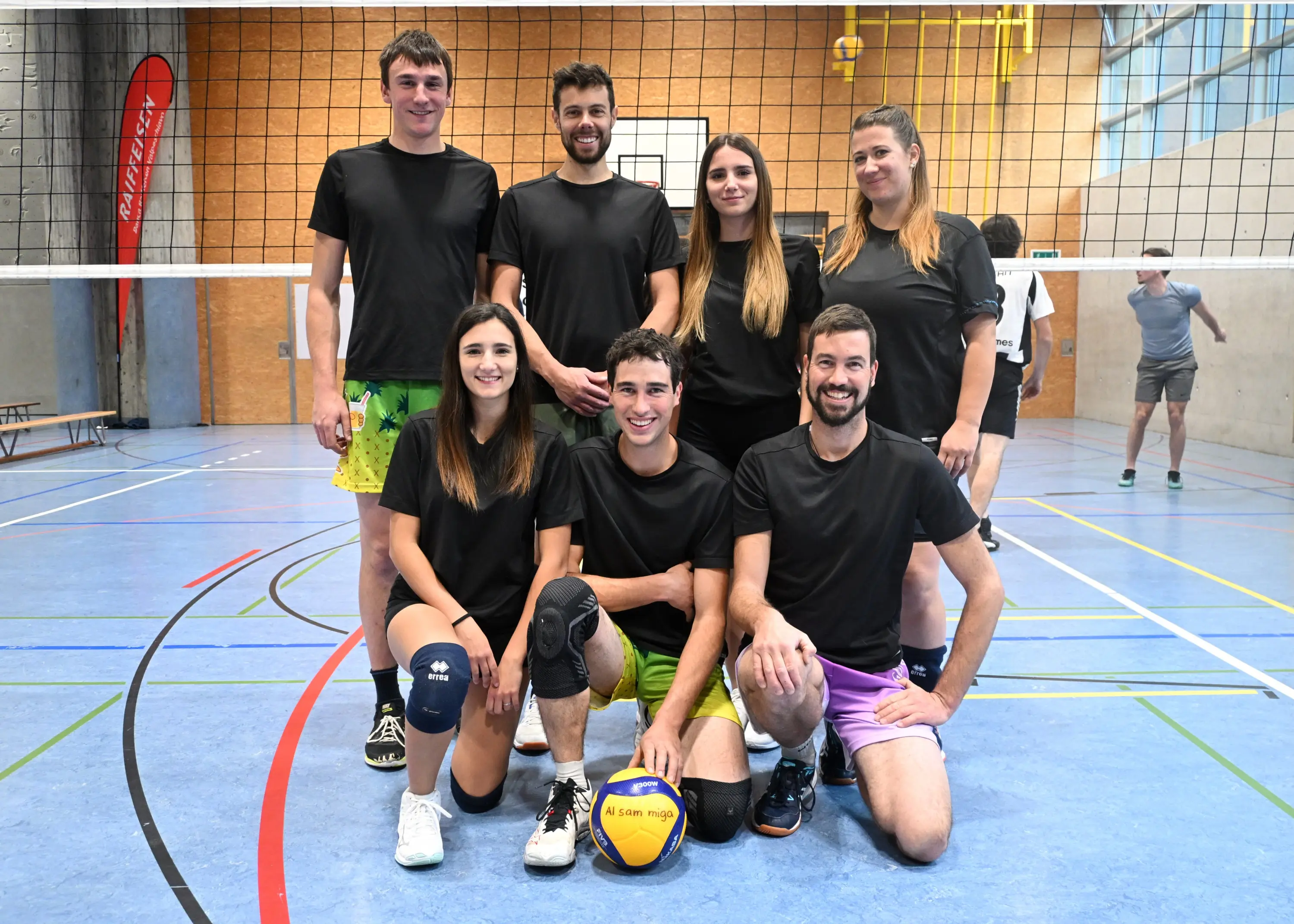 Un gruppo di otto persone sorridenti in abbigliamento sportivo nero, posano in un campo di pallavolo. In primo piano, un ragazzo tiene un pallone da pallavolo e si inginocchia, mentre gli altri sono in piedi dietro di lui. Sul