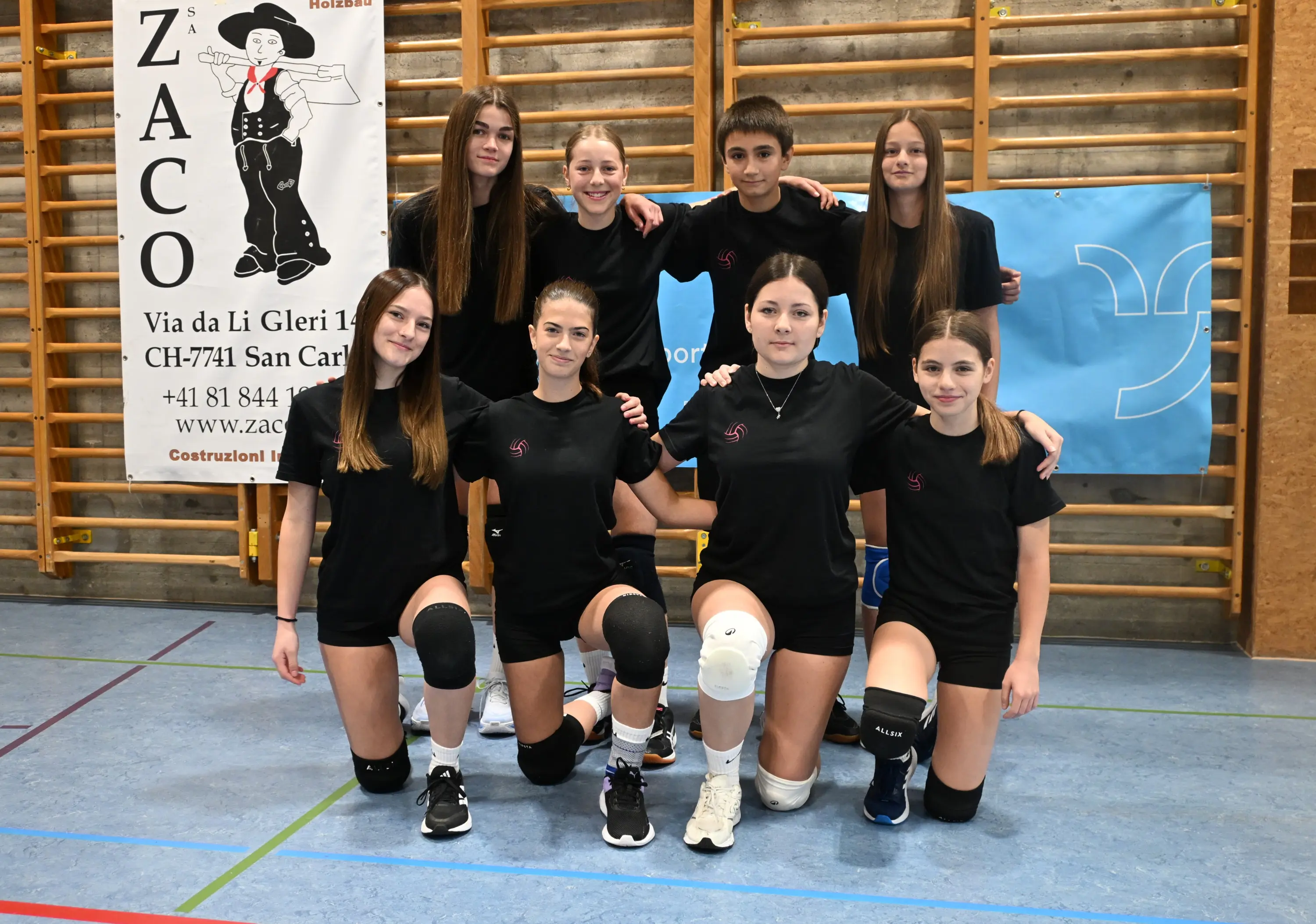 Gruppo di otto giovani atlete di pallavolo, vestite con magliette nere, posano insieme in una palestra. Alcune sono inginocchiate, altre in piedi, sorridono verso la fotocamera. Sfondo con pannelli pubblicitari e attrezzature