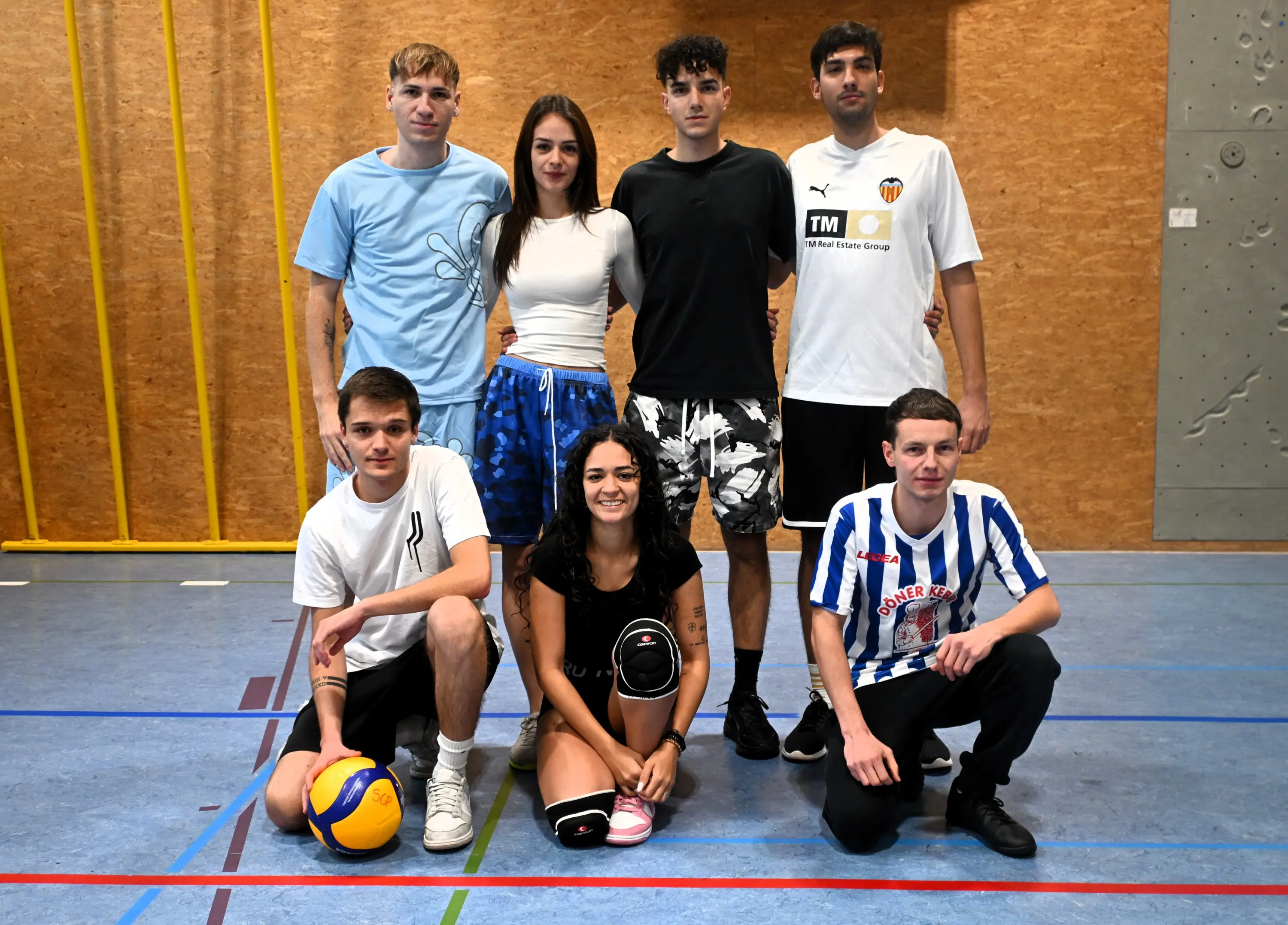 Un gruppo di sette giovani in una palestra, posando insieme. Due di loro indossano maglie sportive e si trovano in piedi, mentre gli altri sono seduti in prima fila. Alcuni hanno palloni da sport e ci sono diverse espressioni di divertimento e amicizia.