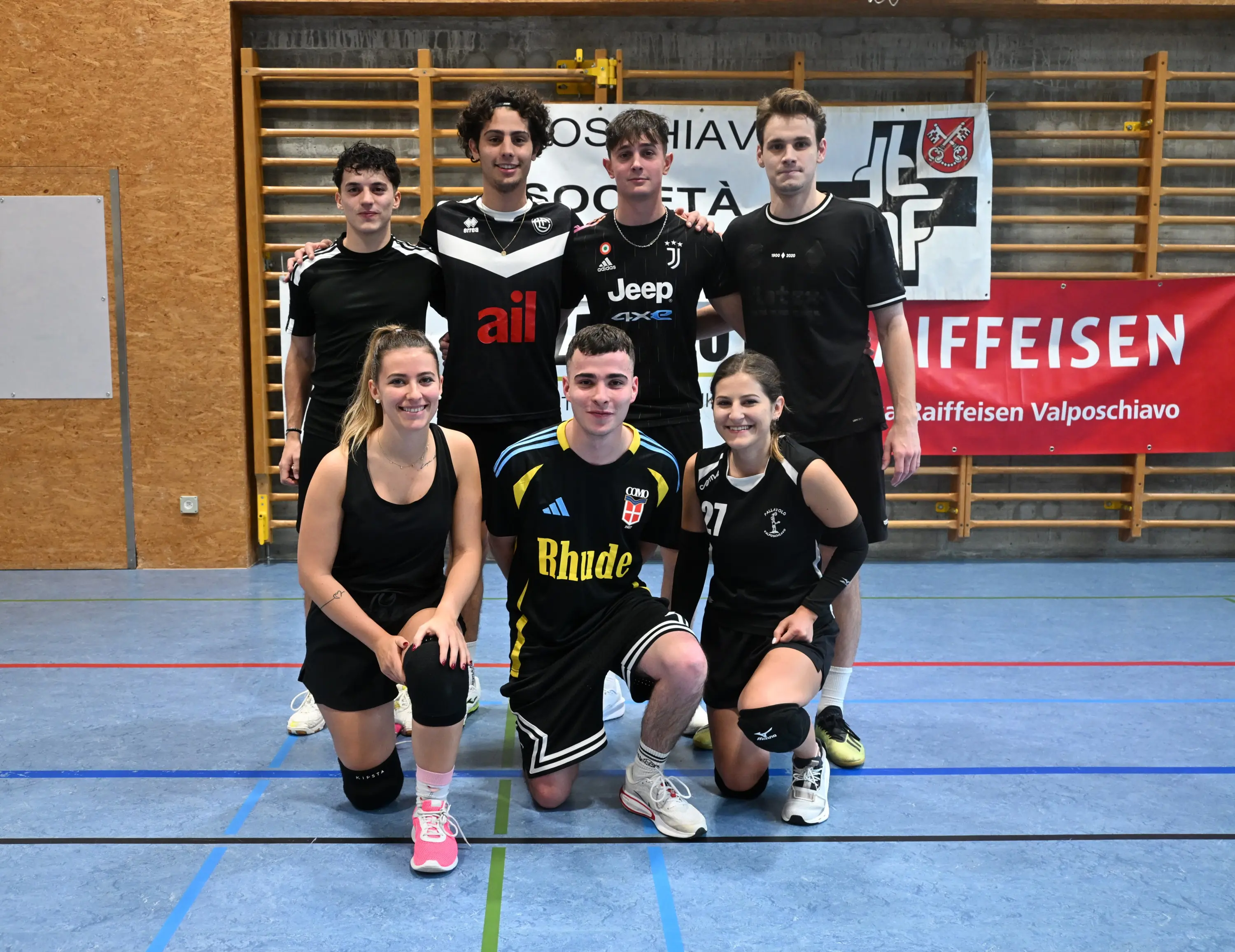 Un gruppo di sette giovani atleti in posa, indossando uniformi sportive, nella palestra di una scuola. Sullo sfondo si vedono banner e sponsor.