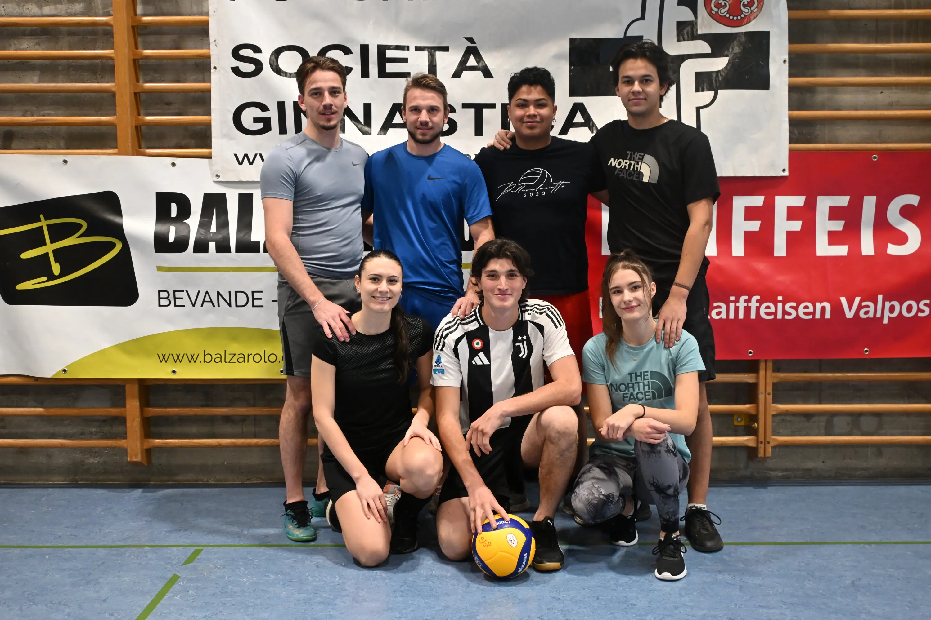 Gruppo di sei persone sorridenti in una palestra, con due uomini in piedi e quattro persone, due uomini e due donne, sedute in prima fila. Sullo sfondo ci sono banner pubblicitari e il logo di una società sportiva.