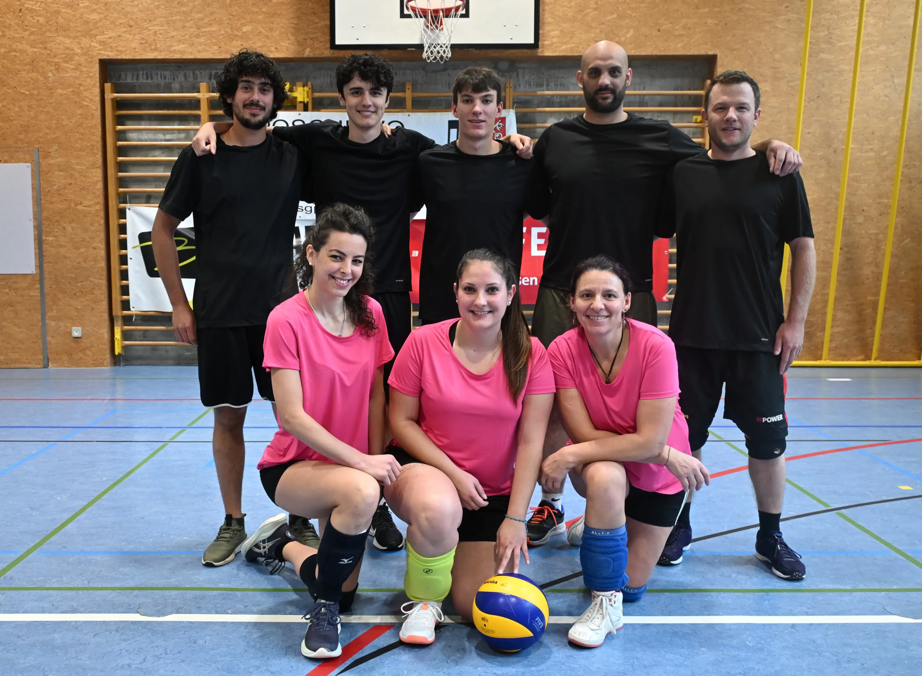 Gruppo di otto persone in una palestra, sorridenti e vestite in magliette nere e rosa. Tre uomini in piedi sullo sfondo e cinque donne e uomini in prima fila, alcuni indossano ginocchiere. Un pallone da pallavolo è posizionato