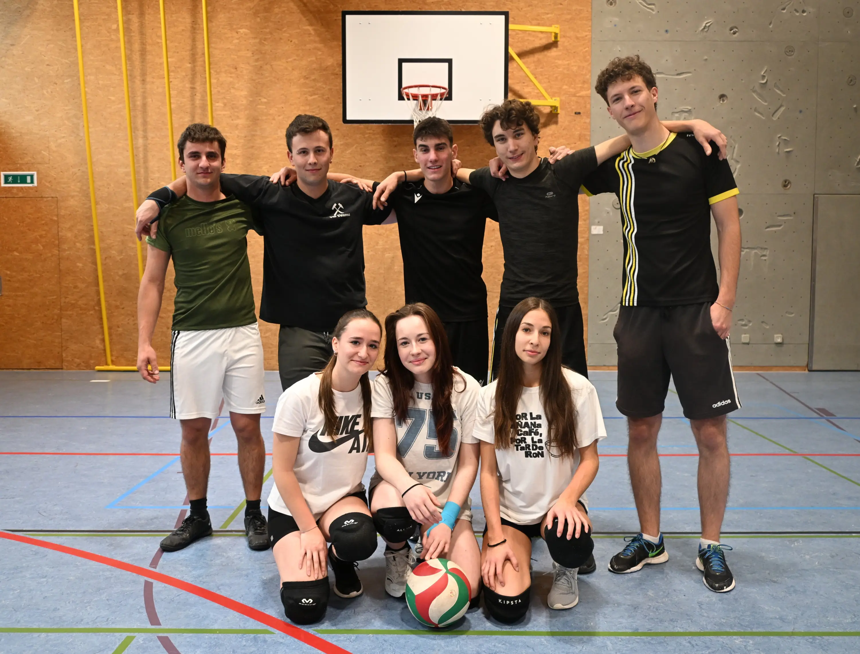 Gruppo di otto giovani atleti in una palestra, sorridenti e posizionati in due file. Due ragazze si trovano in prima fila con una palla da pallavolo, mentre sei ragazzi sono dietro di loro. Sullo sfondo si vede un campo da pallav