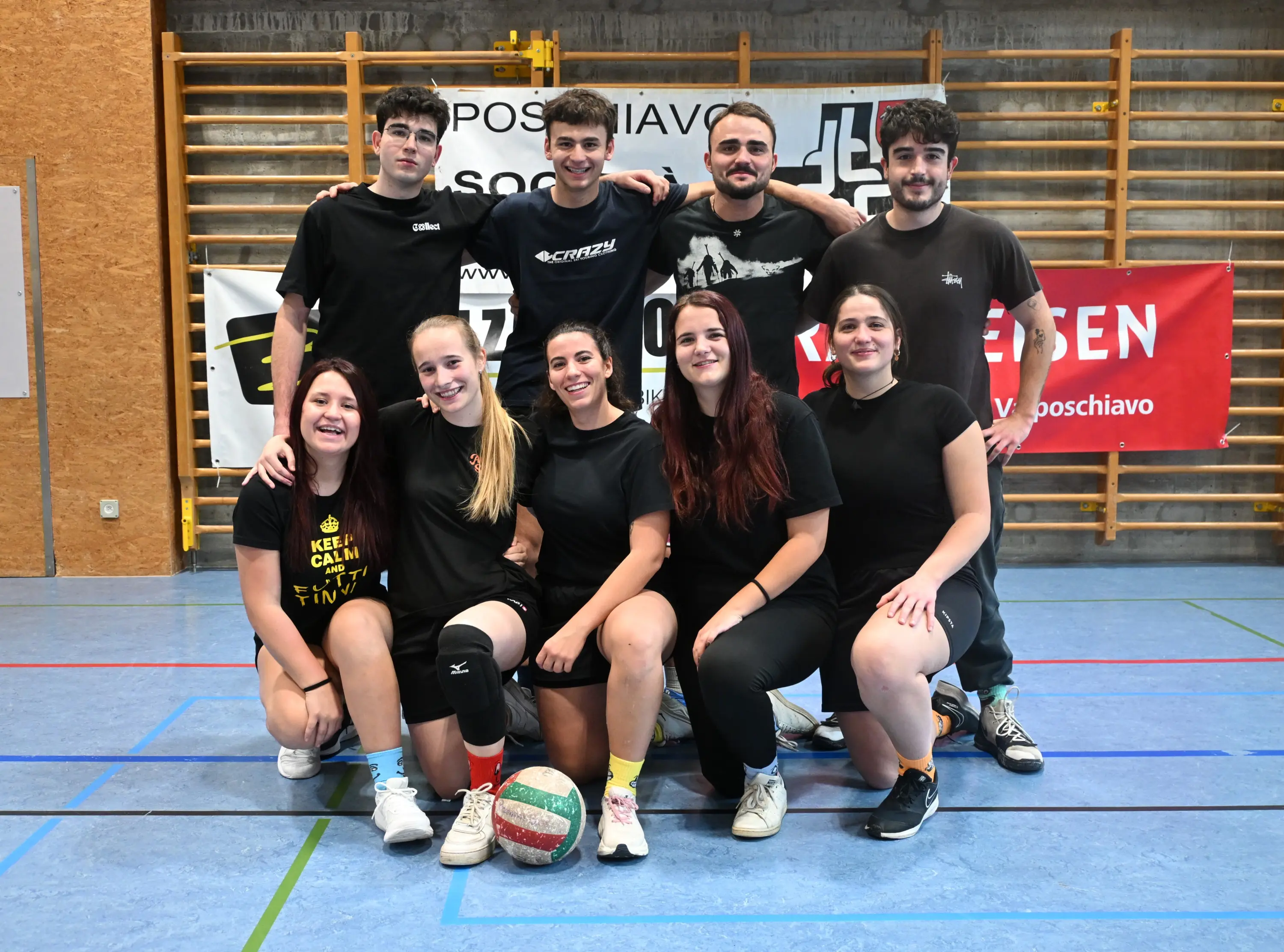 Un gruppo di otto giovani sportivi in t-shirt nere sorride verso la fotocamera. Alcuni sono in posizione accovacciata, altri in piedi, con una palla da pallavolo davanti a loro. Sullo sfondo, ci sono attrezzature sportive e dei banner