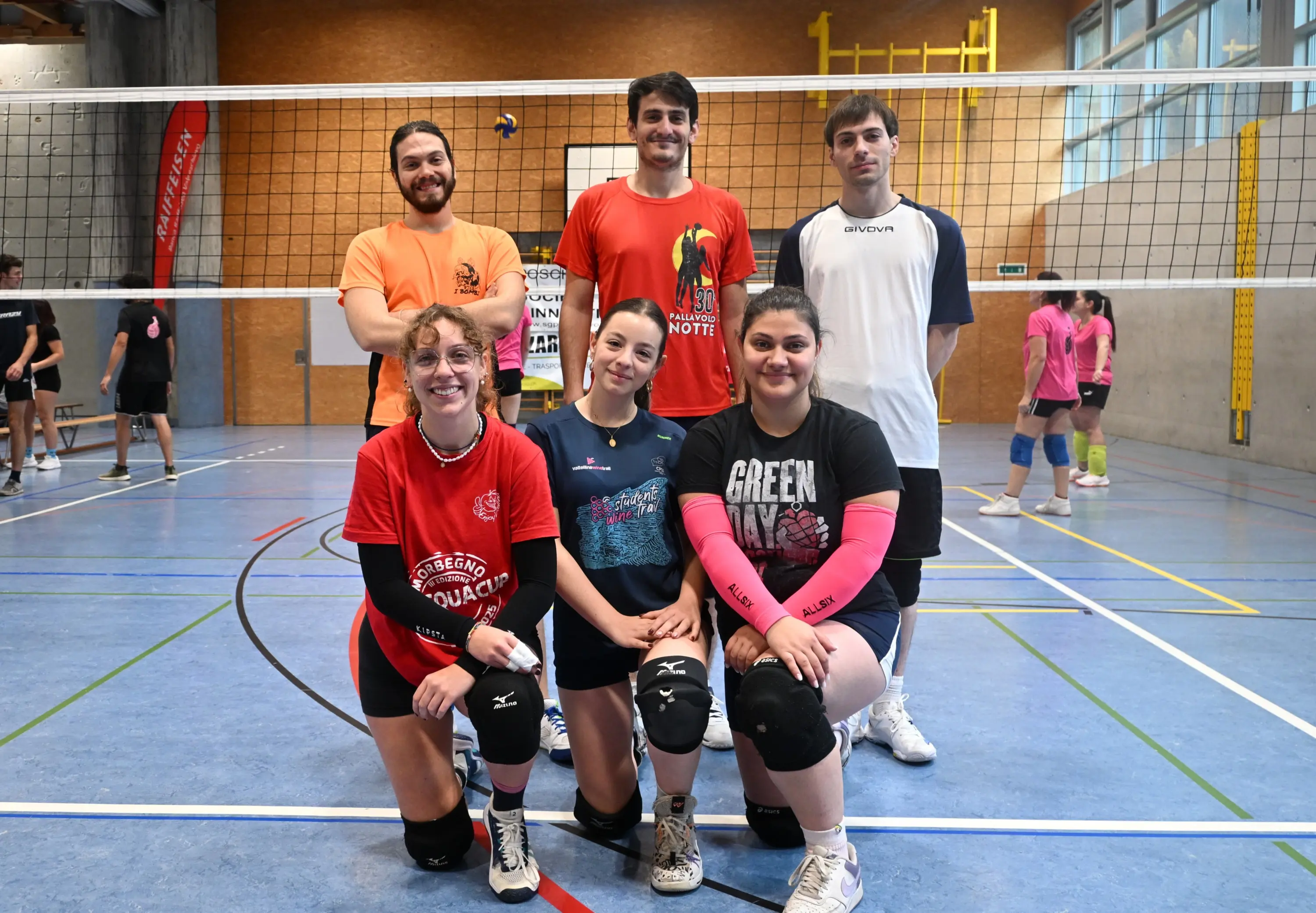 Gruppo di sei persone in una palestra di pallavolo, due in piedi sullo sfondo e quattro accovacciate in primo piano. Indossano magliette colorate e shorts sportivi. La rete di pallavolo si trova dietro di loro. Altri giocatori