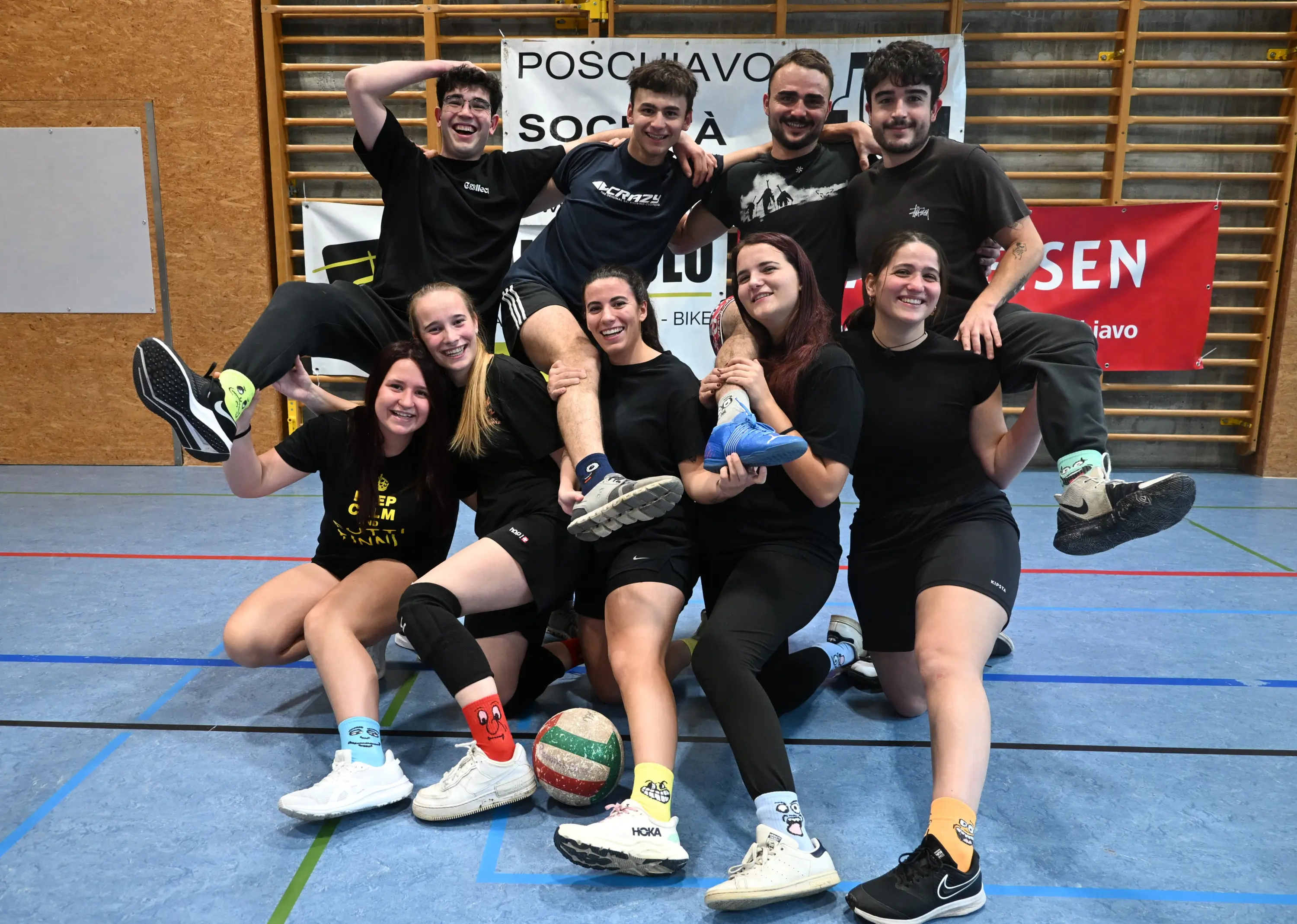 Un gruppo di dieci giovani in maglietta nera sorridono, posando per una foto. Alcuni si tengono in piedi, altri sono accovacciati, con un pallone da pallavolo e calze multicolori. Sullo sfondo ci sono banner e