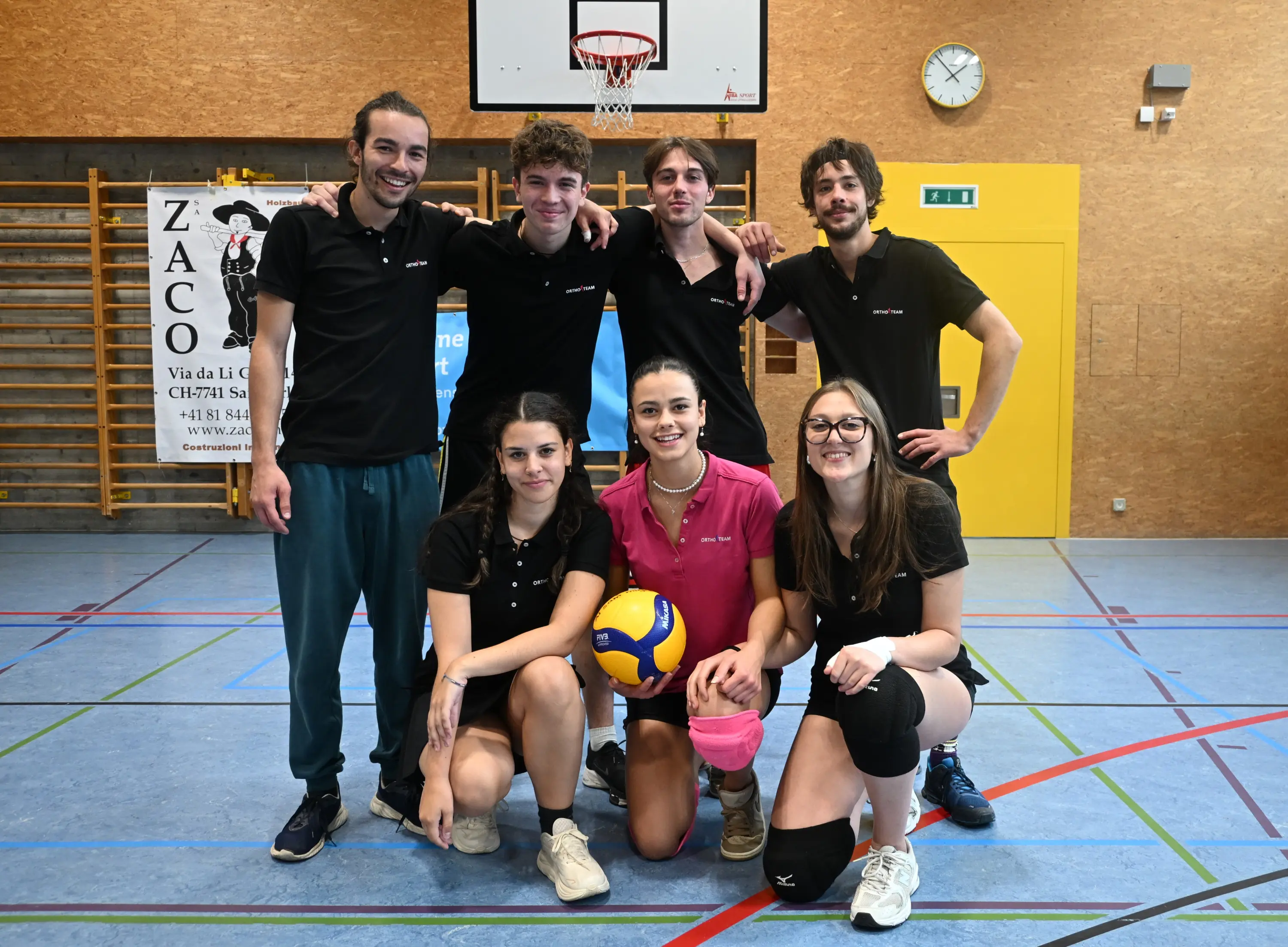 Un gruppo di sette giovani atleti in una palestra di volley, sorridenti e in abbigliamento sportivo, posano per una foto. Alcuni tengono in mano un pallone da volley. Sullo sfondo ci sono attrezzature sportive e un canestro.
