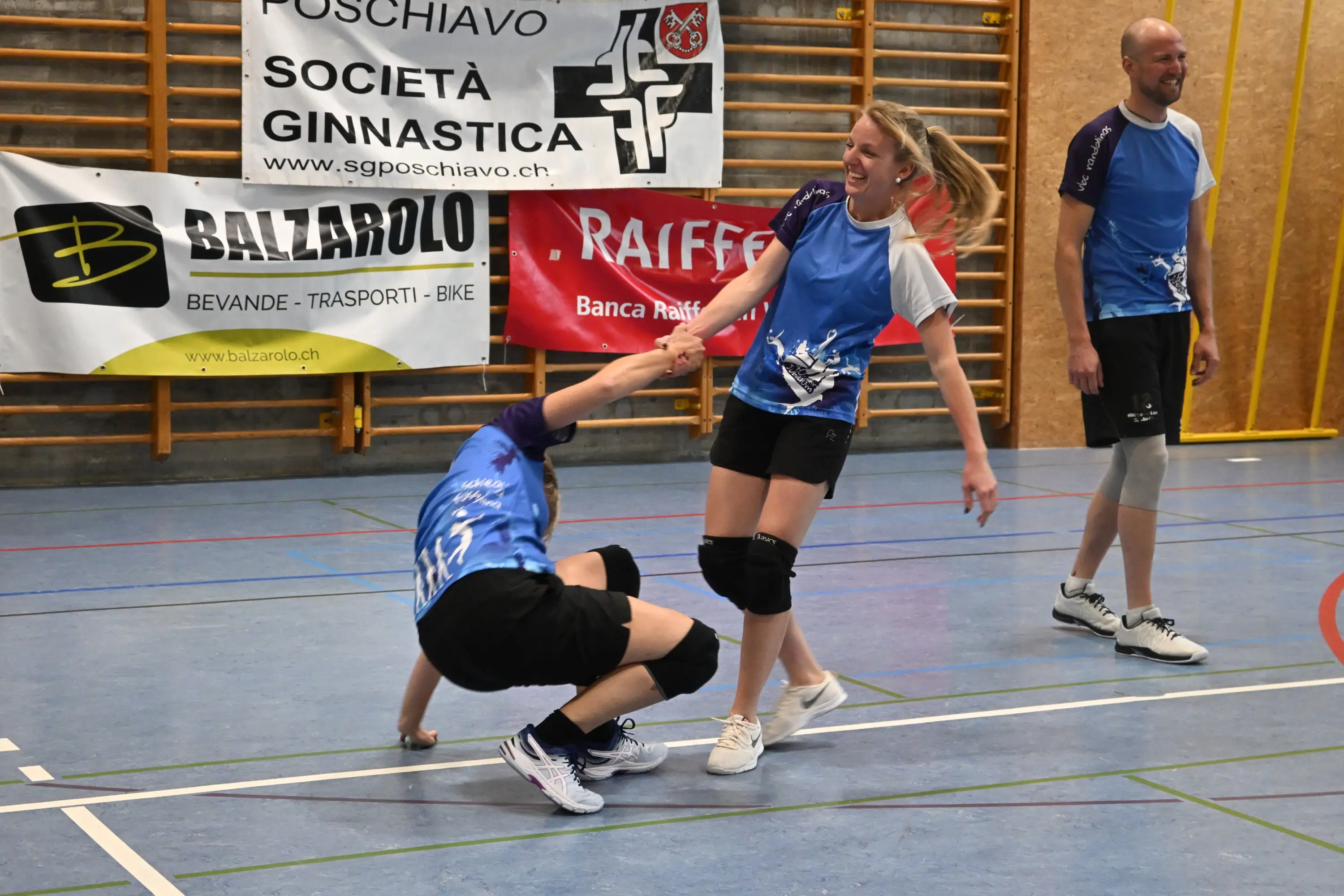 Due giocatori di pallavolo si divertono in campo. Una giocatrice aiuta un compagno che è caduto, entrambi sorridono. Sullo sfondo ci sono striscioni pubblicitari.
