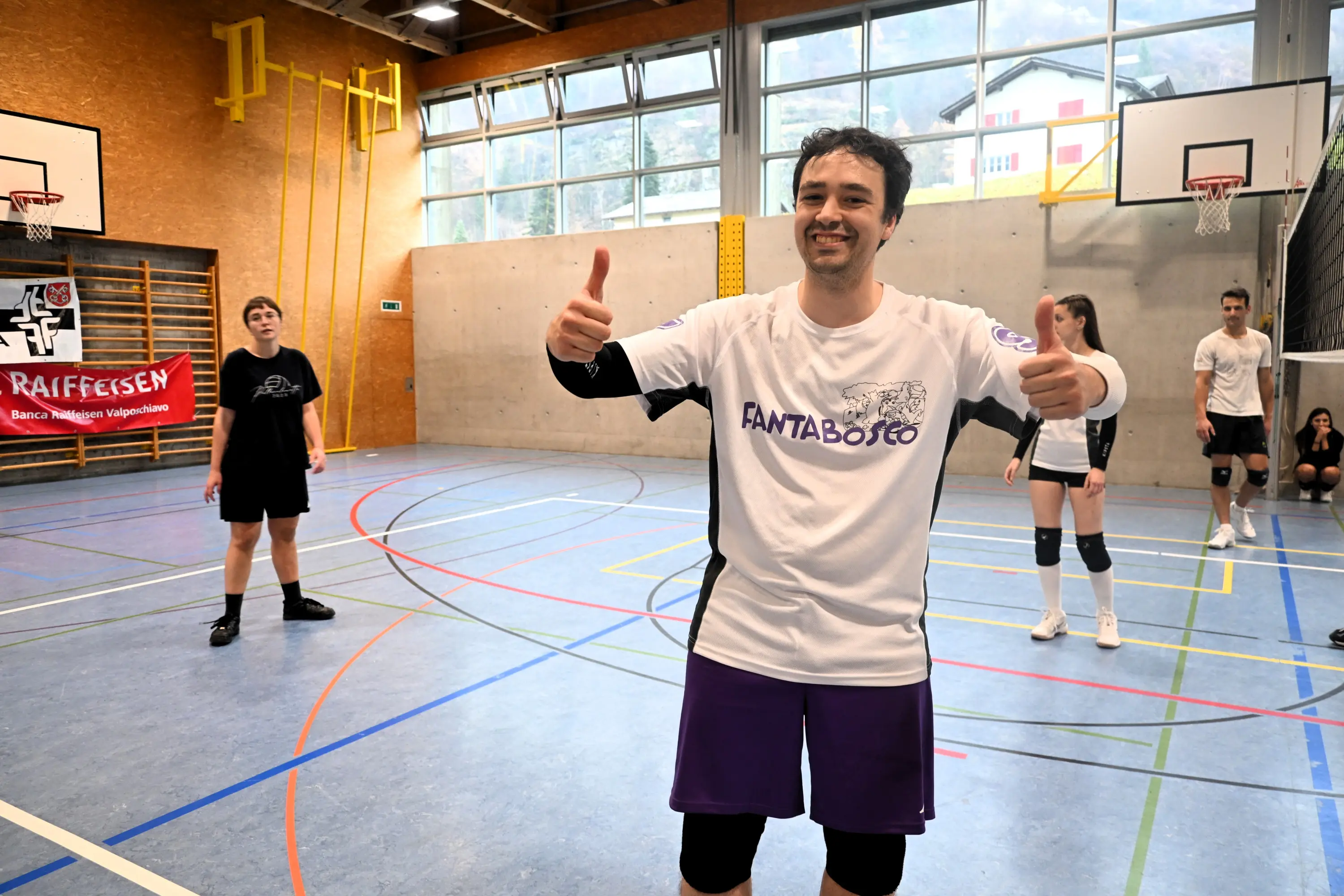 Giocatore di pallavolo in una palestra, sorridente e con il pollice in su, indossa una maglietta bianca con "FANTABO" e pantaloni viola. Sullo sfondo, due altri giocatori sono in azione, mentre una bandiera ro