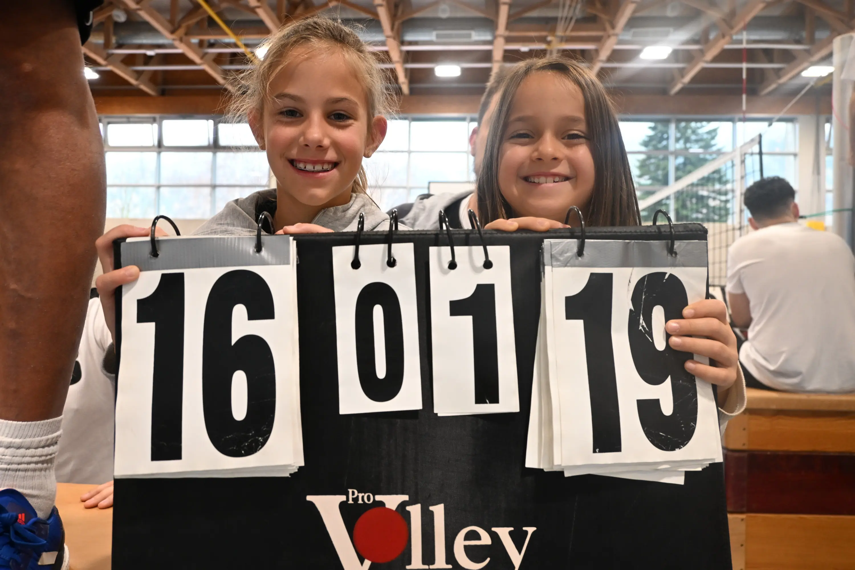 Due ragazze sorridenti tengono in mano un tabellone di punteggio, che mostra i numeri "16", "01" e "19". Sono in un ambiente sportivo, probabilmente una palestra.