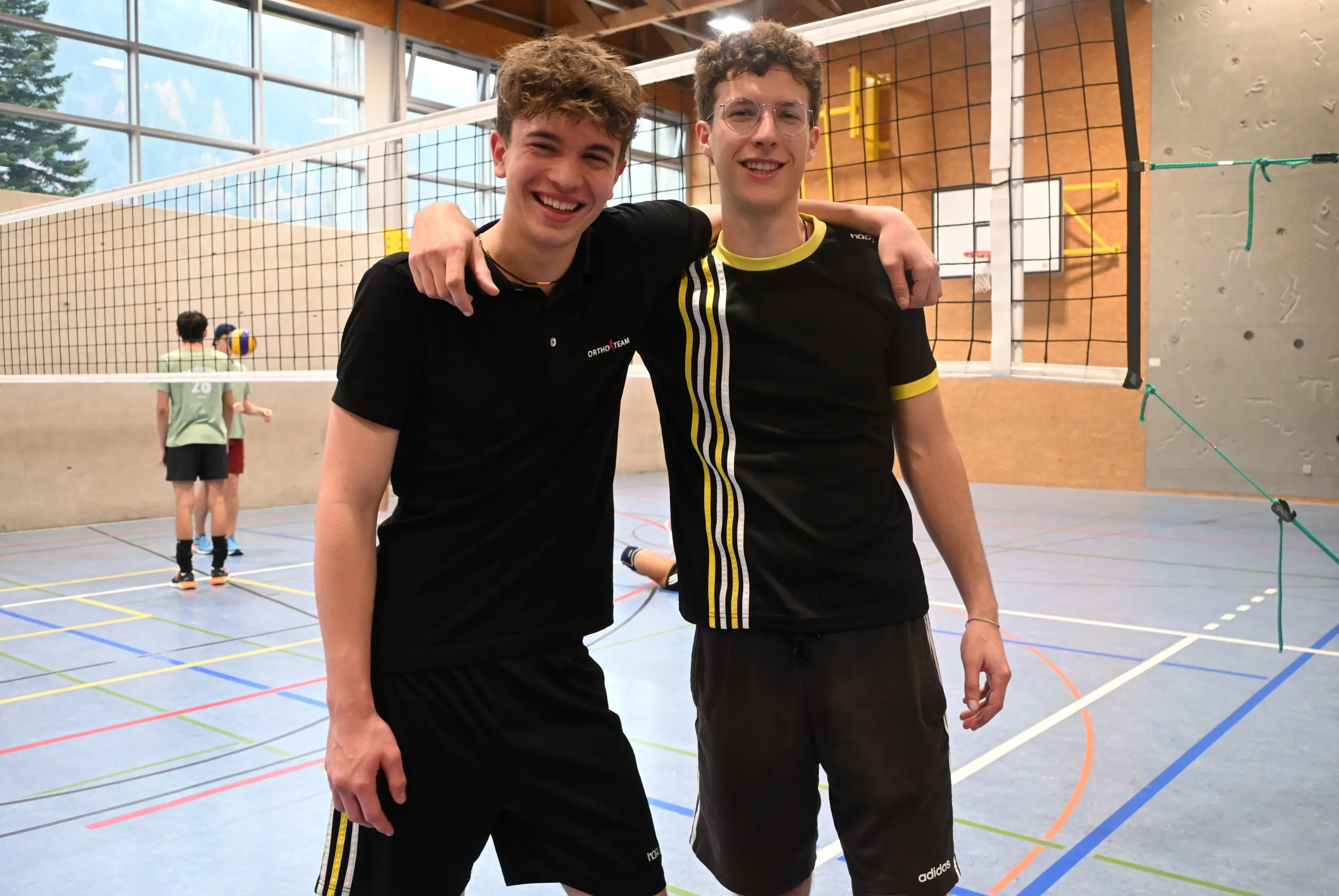 Due giovani sorridenti in maglietta sportiva, abbracciati e posando per una foto in un campo da pallavolo. Sullo sfondo, altri ragazzi stanno giocando.