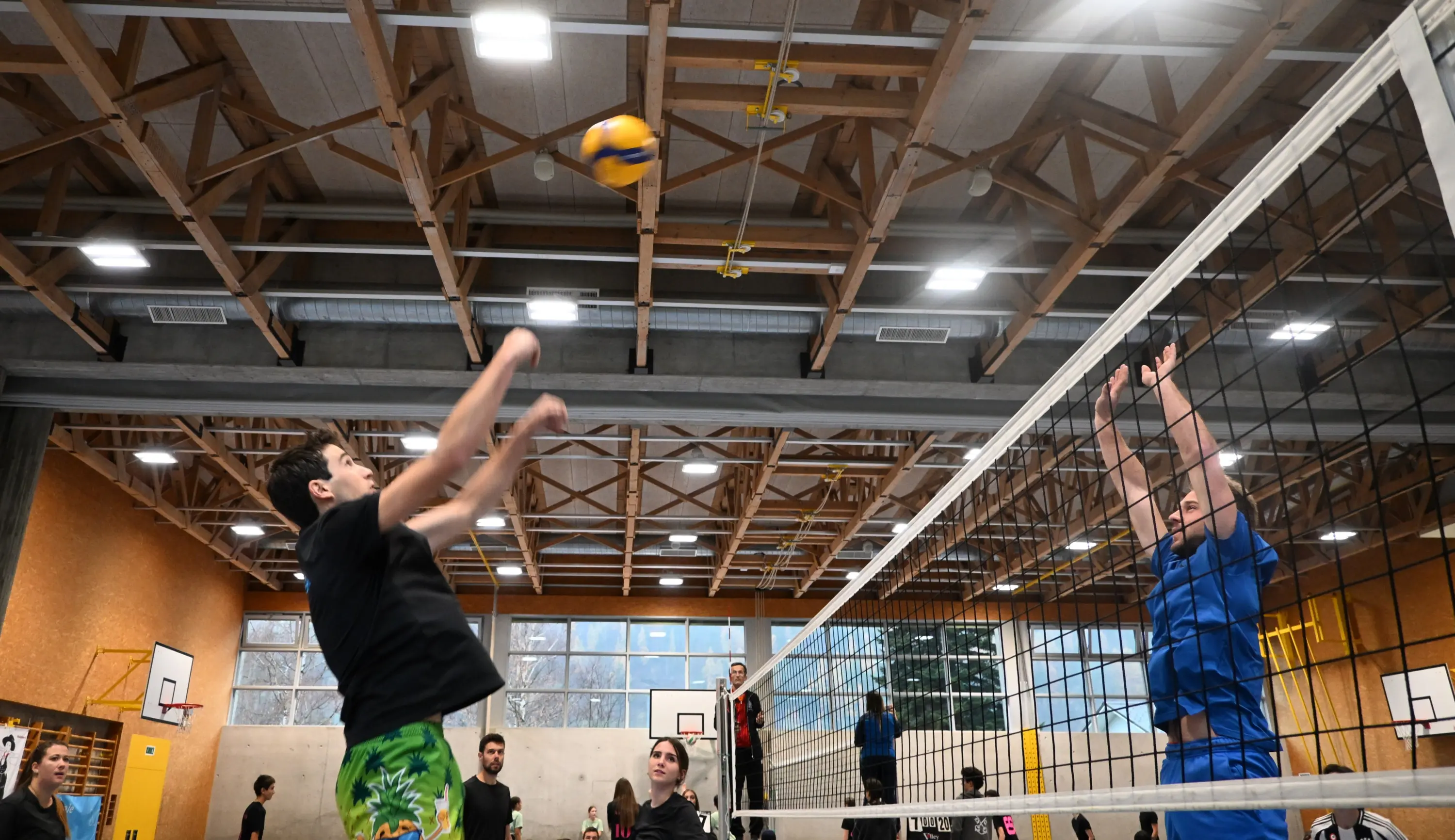 Giocatori di pallavolo che si sfidano in un campo interno, con uno che salta per colpire la palla e l'altro pronto a bloccare. Gli spettatori guardano dal fondo.